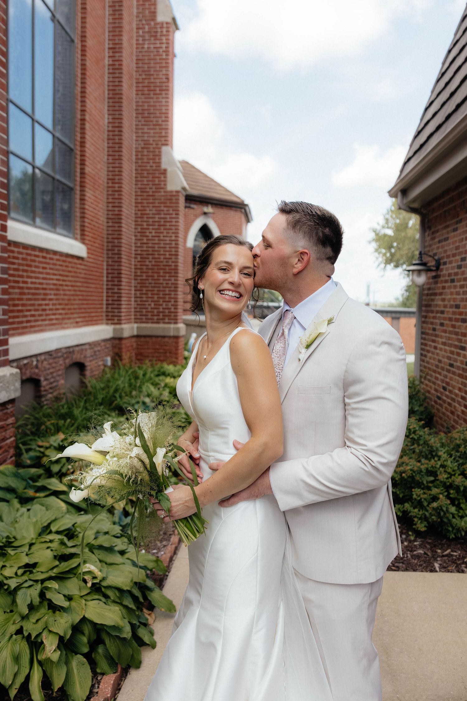 A romantic couple in wedding attire share laughter and embrace while posing together outside a brick building.