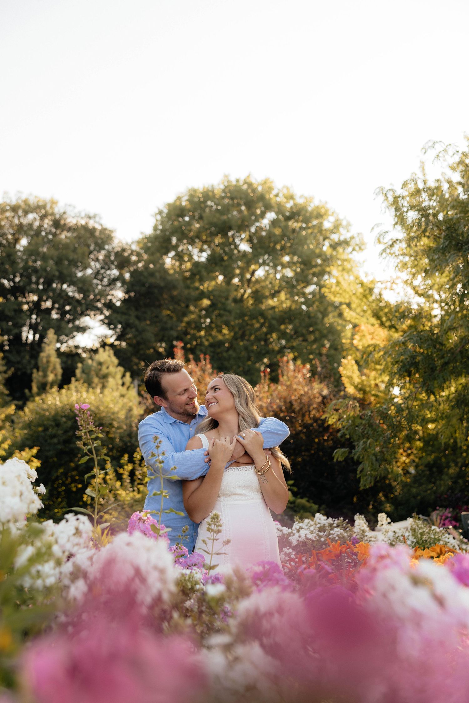 Couple embracing in a colorful garden surrounded by pink, white, and orange flowers with lush green trees at golden hour.