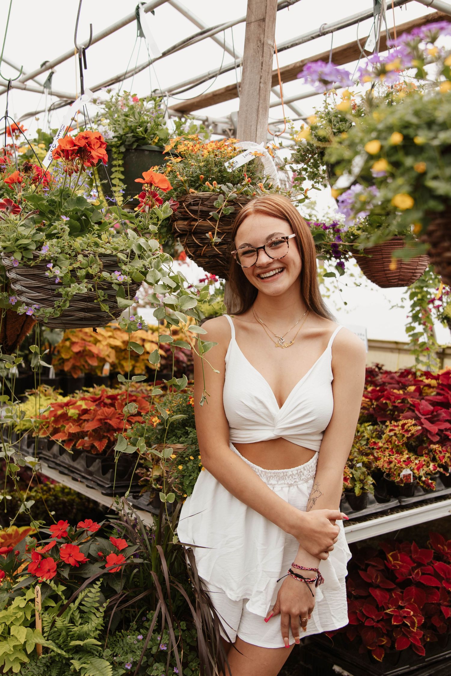 A young high school senior in summer white outfit smiles while surrounded by colorful flowers in a greenhouse.