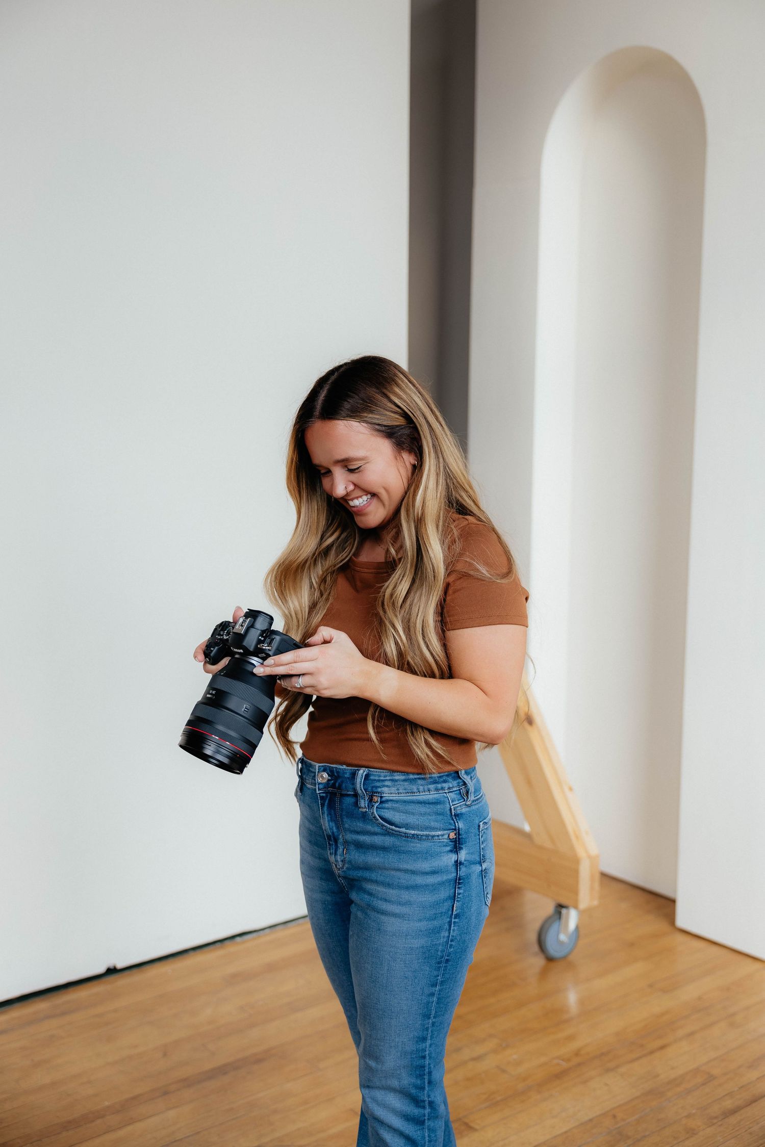 A person in a brown shirt and jeans smiles while looking at the screen of their professional camera in a bright room.