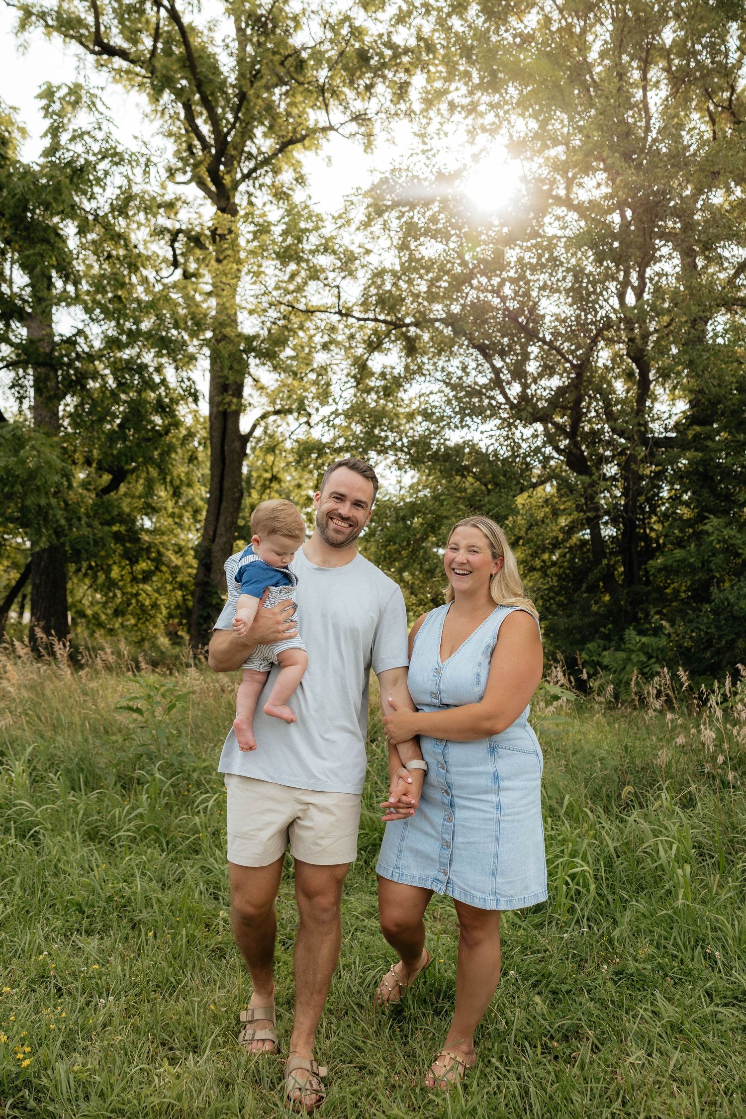 A small family stands together in a sun-dappled grassy field on a warm summer evening.
