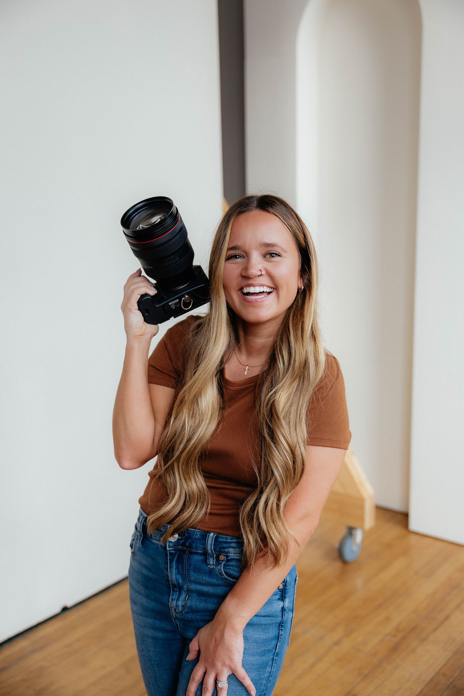Professional photographer in brown shirt and jeans holds camera during indoor studio photoshoot on wooden floor.