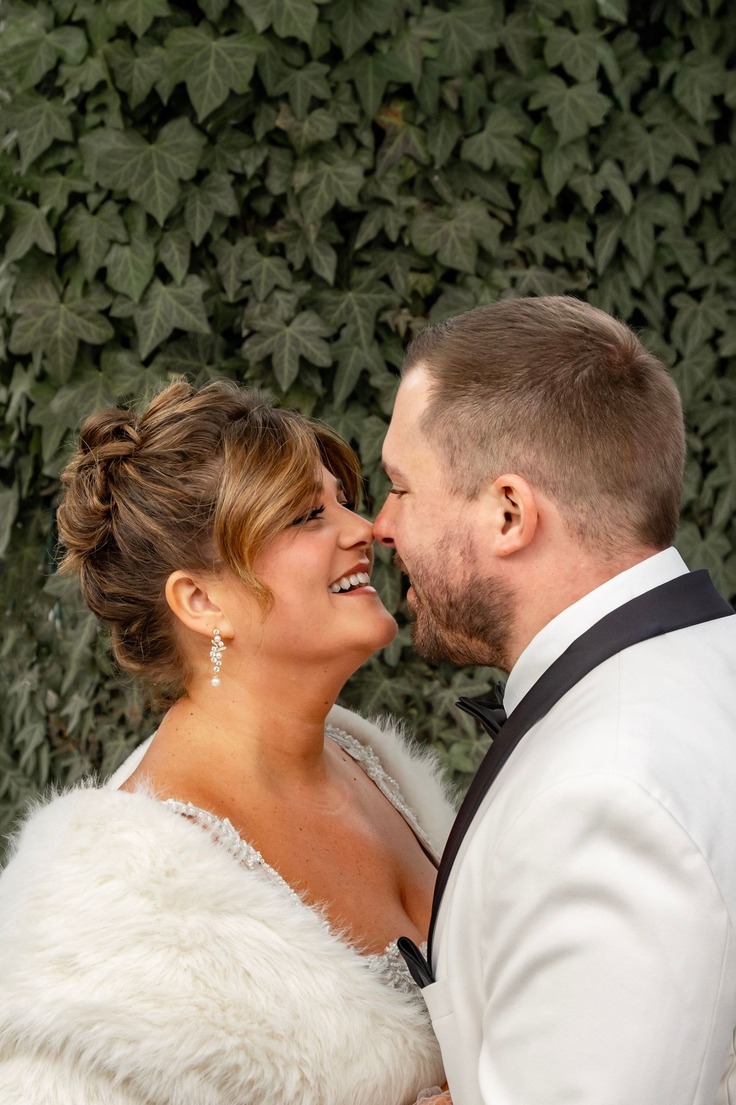 Bride and groom portrait, couple standing in front of a wall of ivy smiling and touching noses.