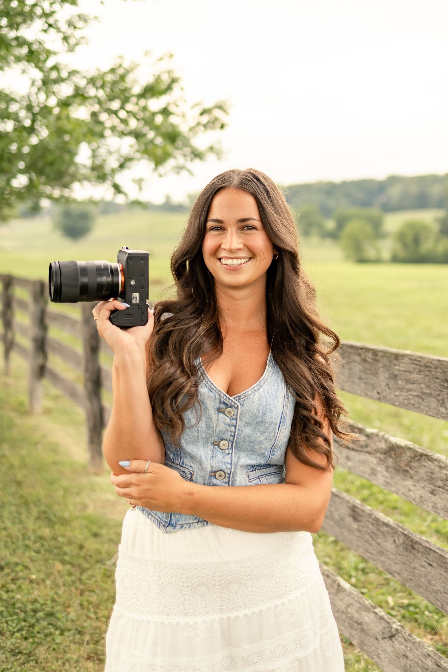 Photographer branding photo, photographer with brown curly hair wearing a denim vest and white skirt holding a camera in front of a field