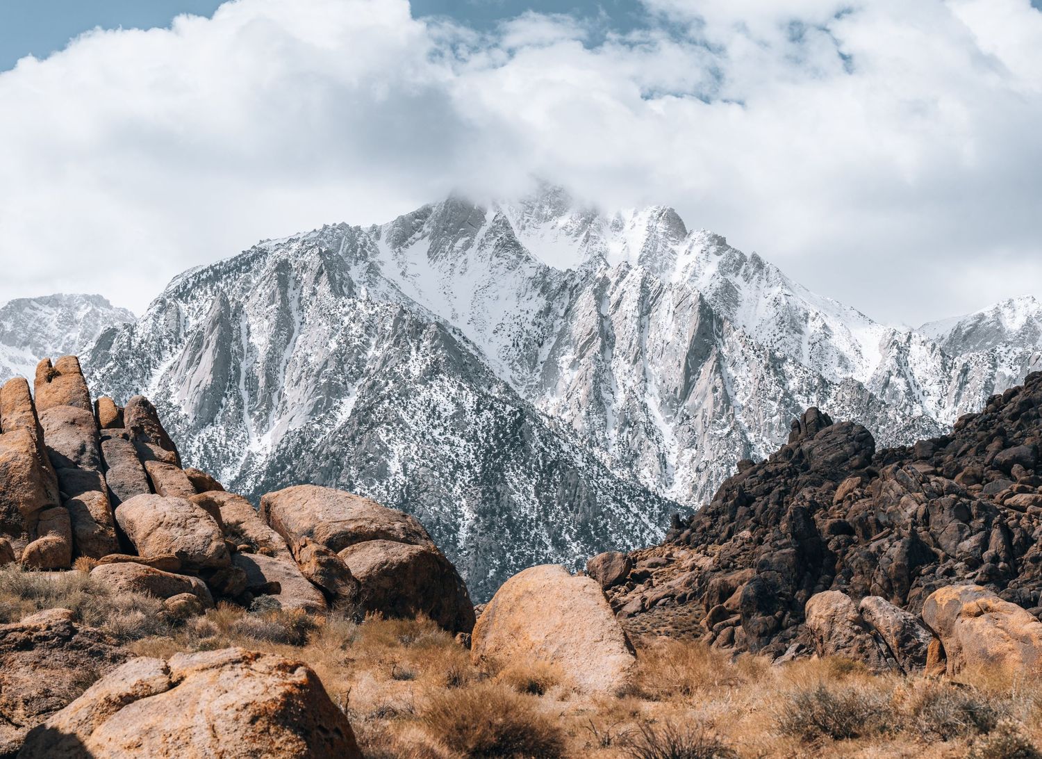 Rocky desert terrain contrasts with snow-covered mountain peaks under cloudy skies.