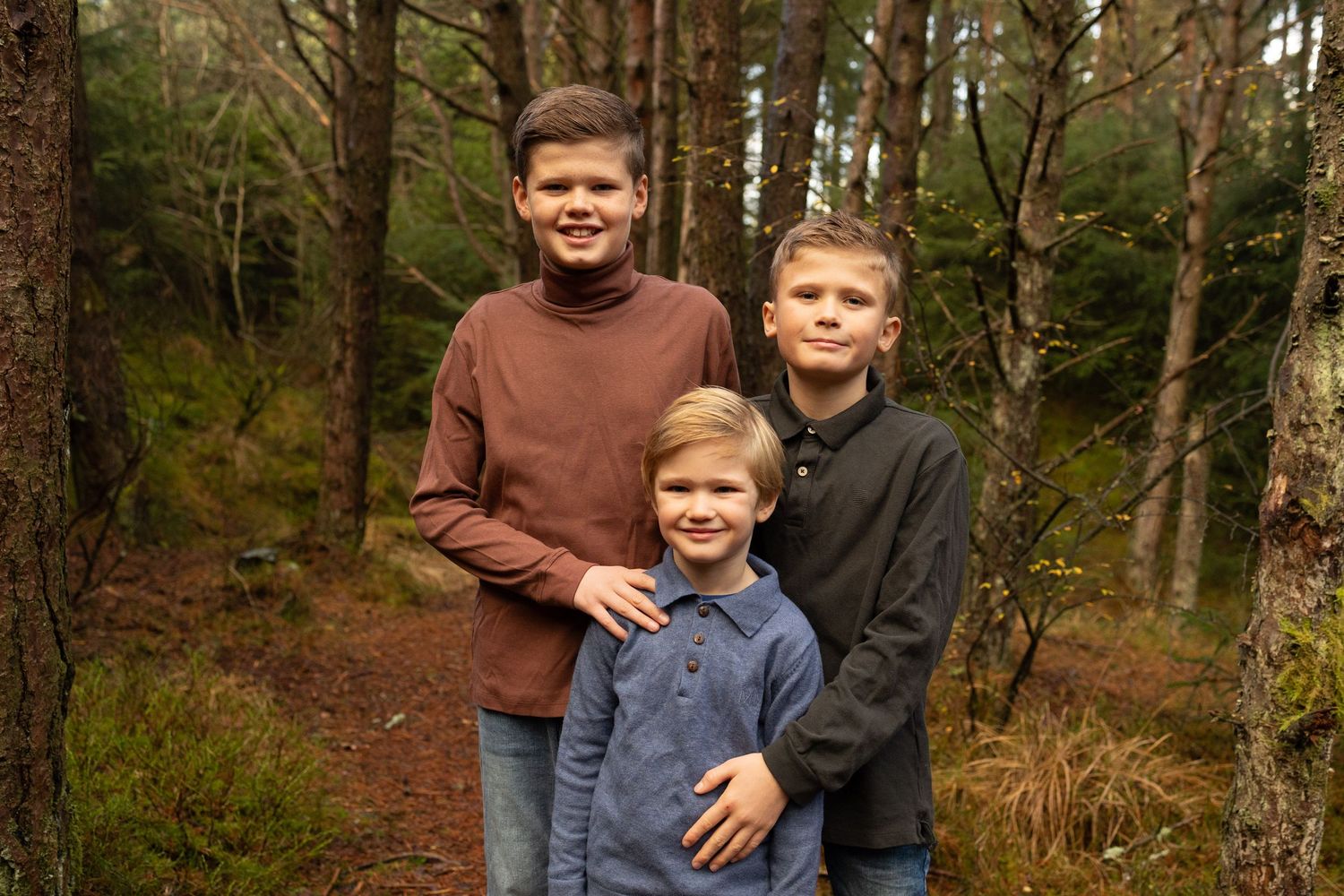Three children stand together on a wooded forest trail wearing casual sweaters on an autumn day.