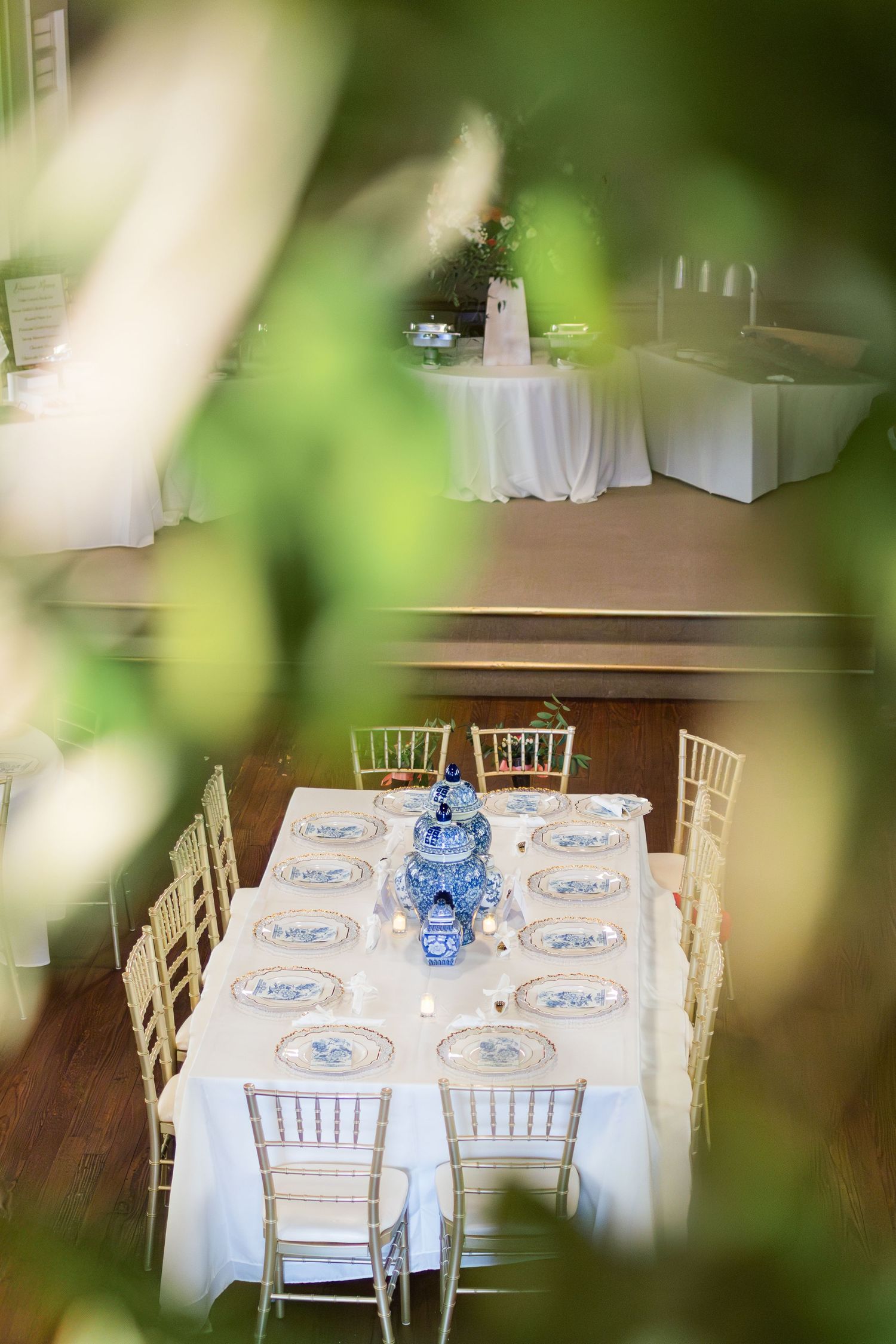 Elegant wedding table settings with blue and white china dishes and a chinoiserie vase centerpiece viewed through foliage.