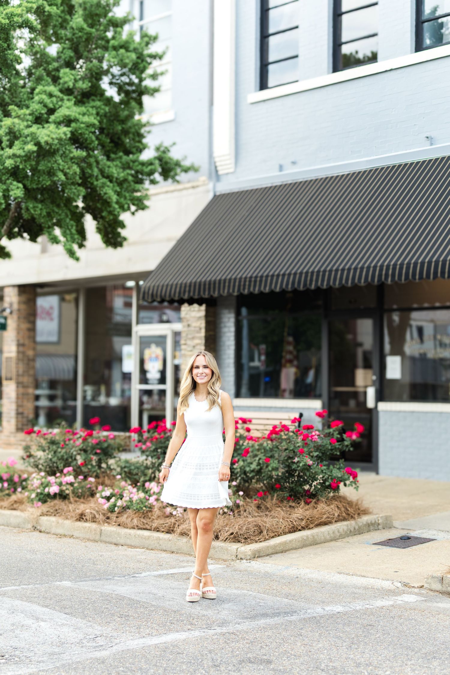 A person in a white dress walks near a storefront with pink flowers and black awnings in an urban setting.