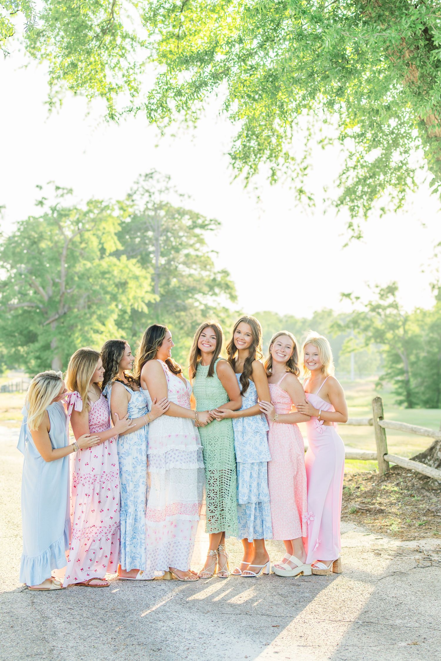 A group in pastel dresses stands together on a sunlit path surrounded by green trees.