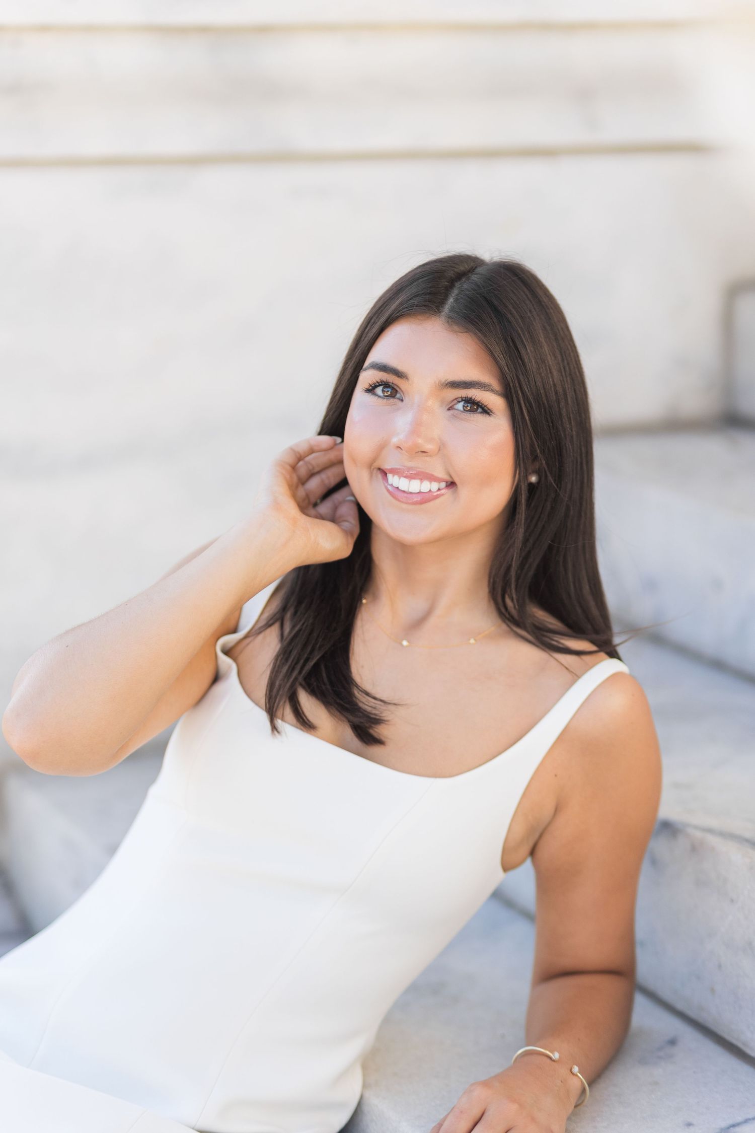 Woman in white tank top poses on concrete steps while smiling and playing with her dark hair in outdoor photoshoot.