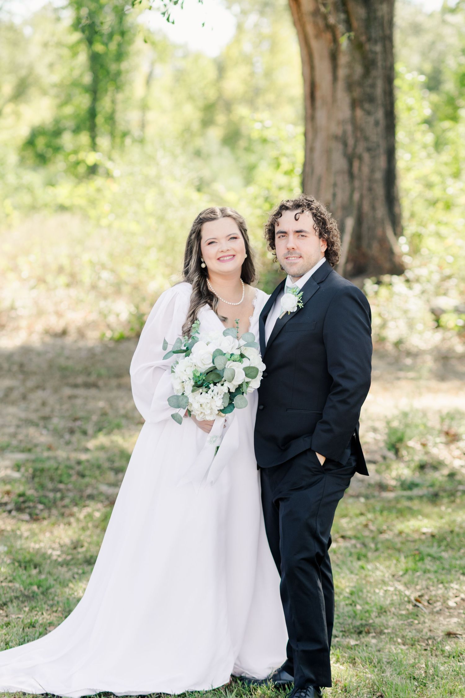 A couple in wedding attire poses together outdoors in a wooded setting with white flowers and greenery.
