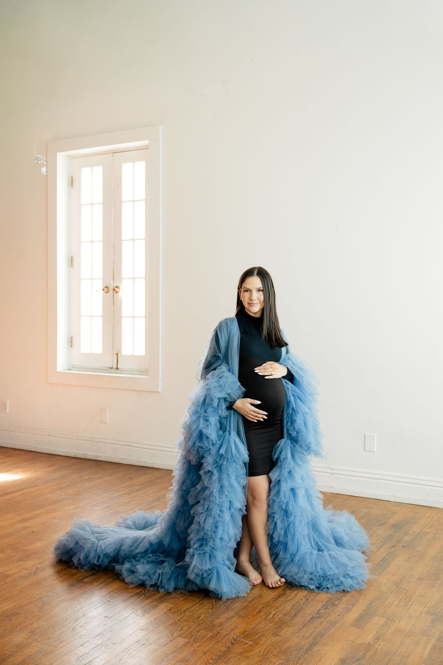 A figure in a black dress and dramatic flowing blue tulle cape poses on wooden floors in a bright white room.