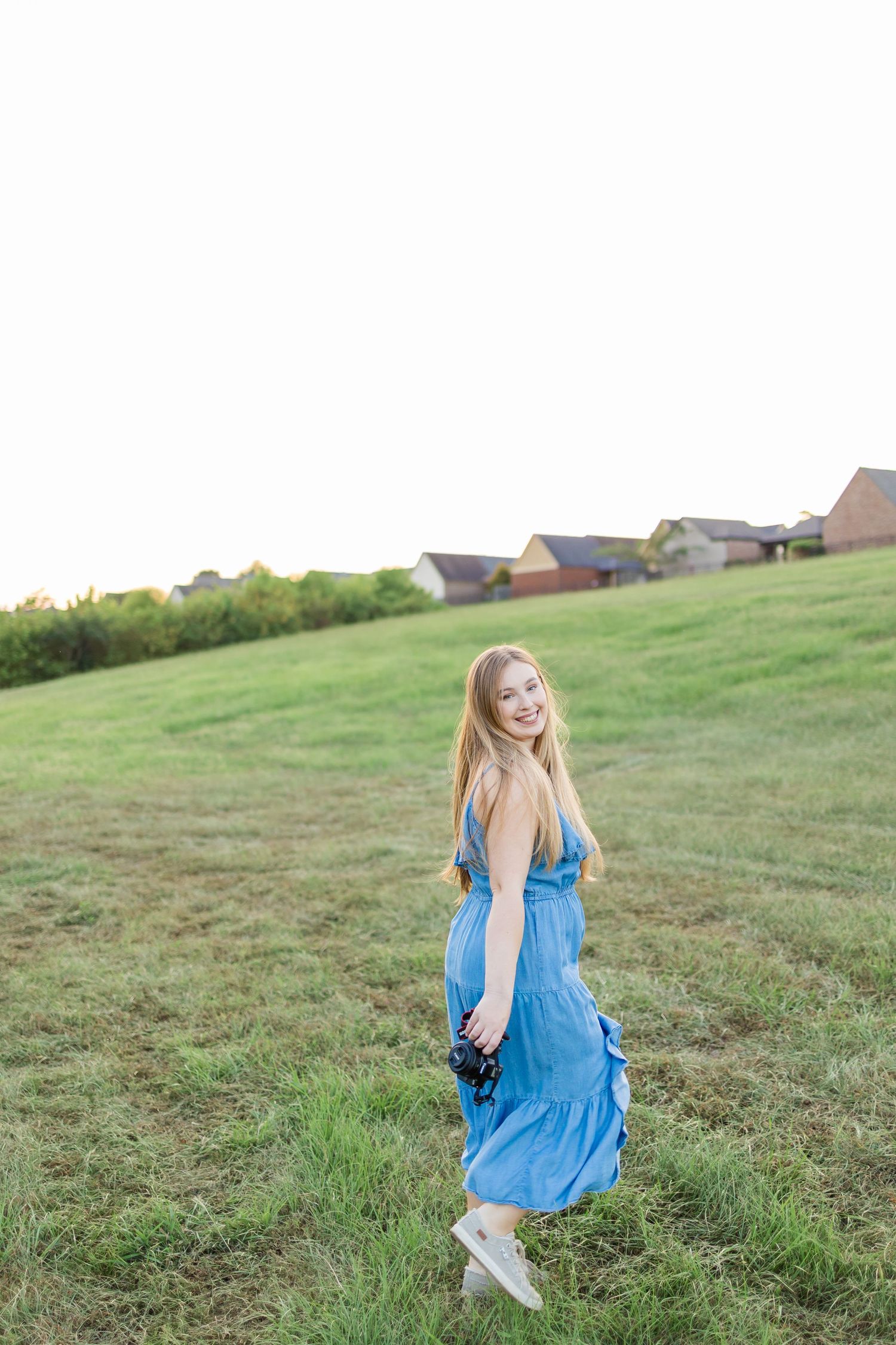 A blue sundress billows in the breeze as someone sits gracefully on a grassy hillside against rolling slopes and houses.
