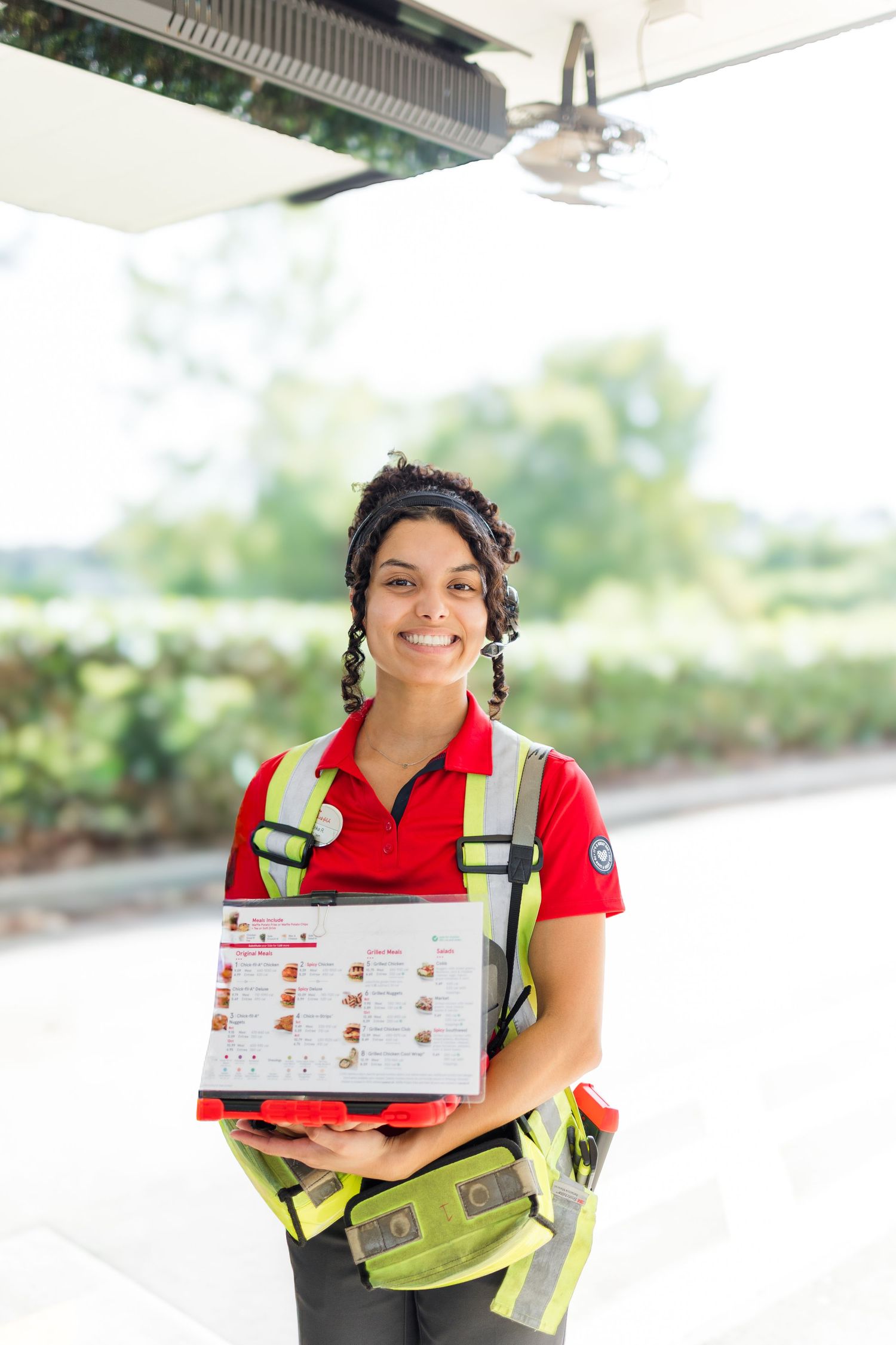 Service worker in red polo shirt and high-visibility vest holds menu or order form while smiling outdoors.