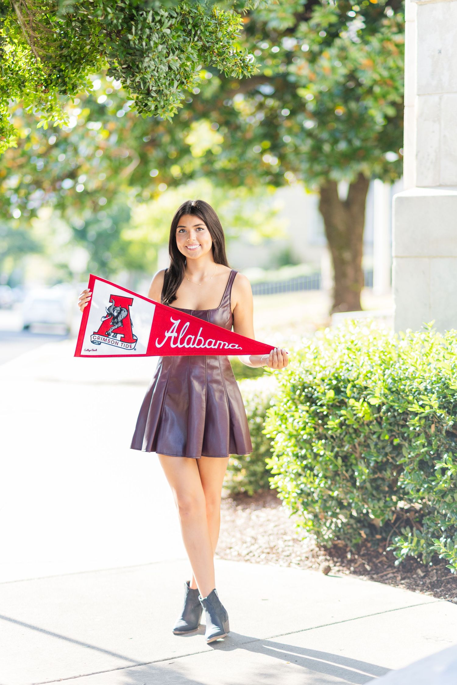 A person in a brown dress and boots poses outdoors holding a red pennant flag near green shrubbery.