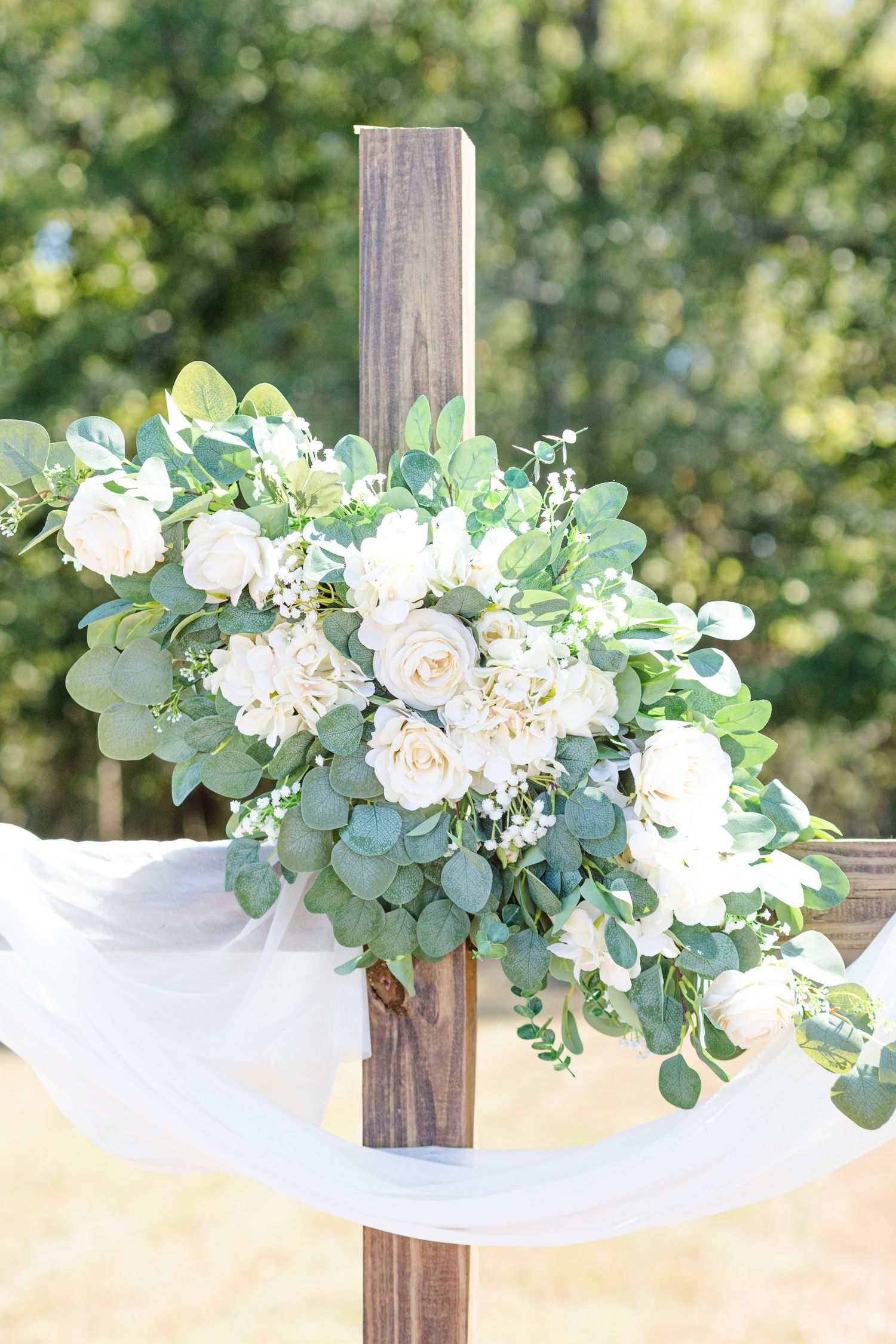 White fabric drapes elegantly across wooden posts adorned with lush eucalyptus and white rose floral arrangements.