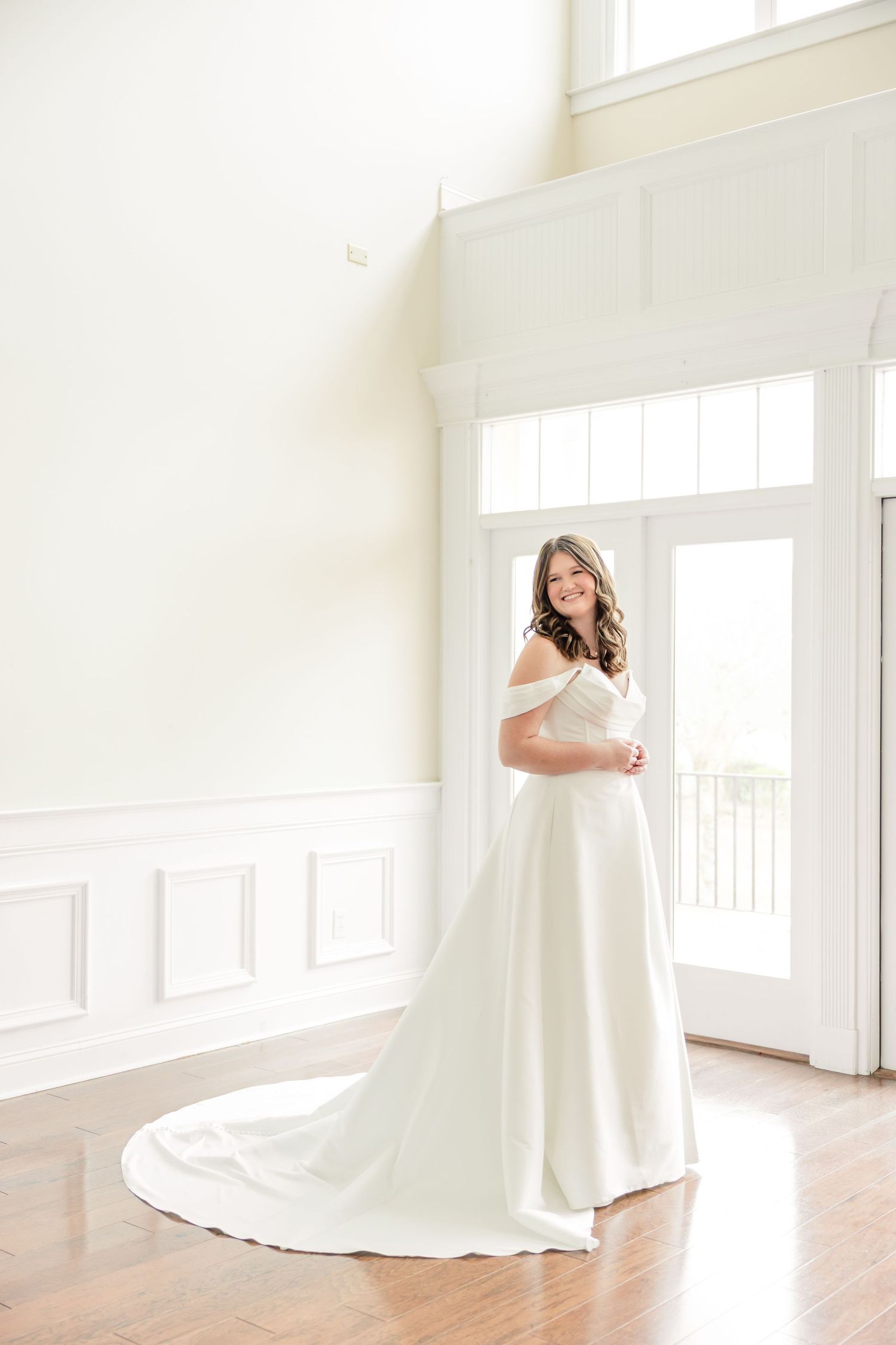 A bride in a flowing white wedding gown twirls gracefully in a bright, sunlit room with wooden floors.