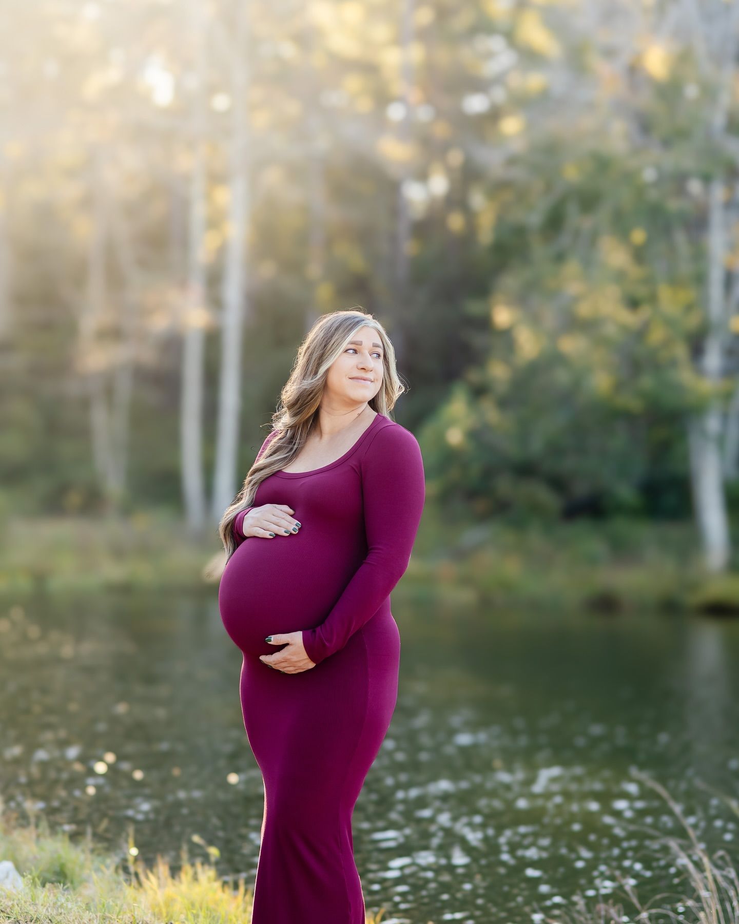 A maternity photo of a pregnant woman in a burgundy dress standing by a lake at sunset.
