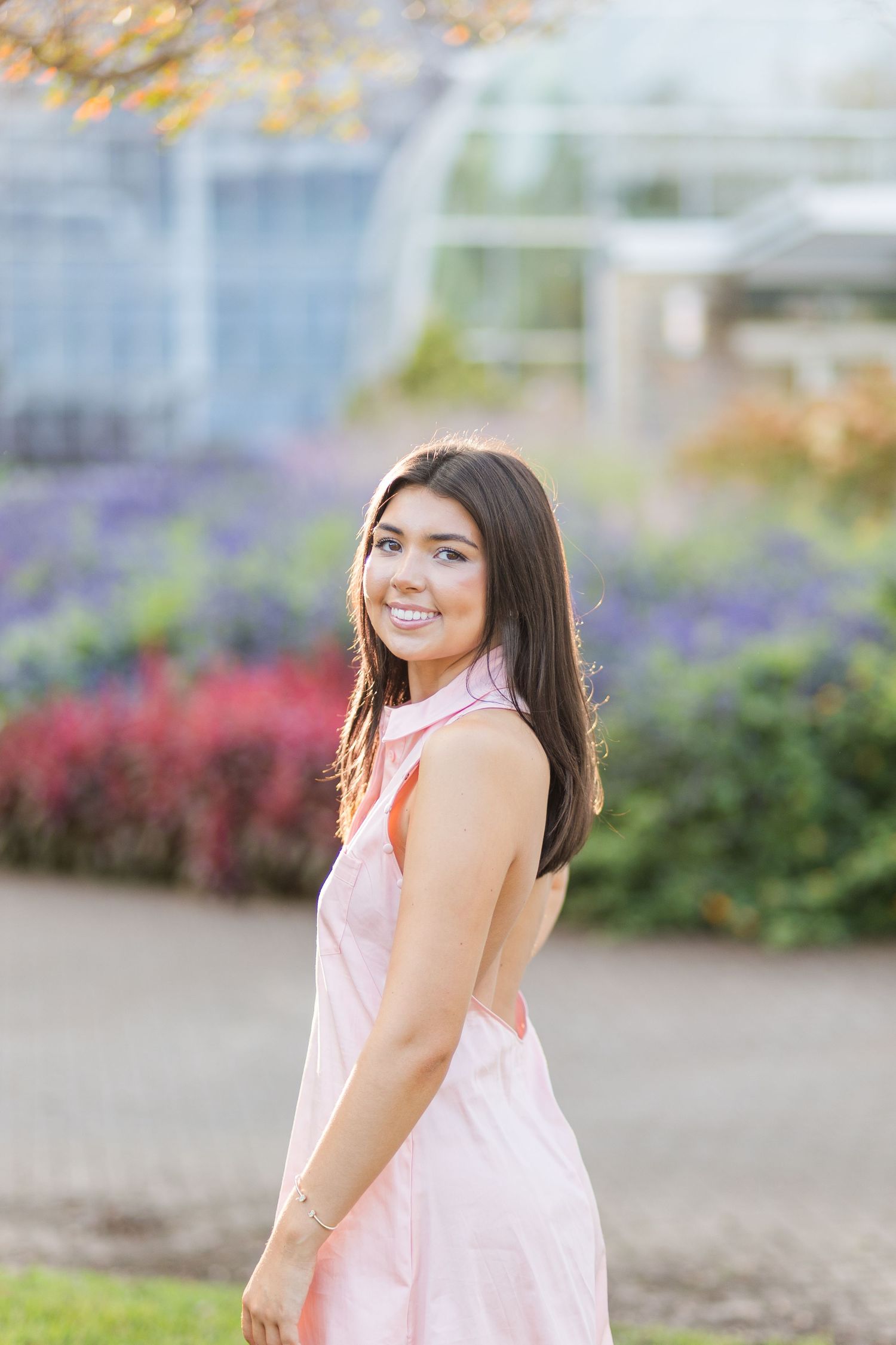 Person in a light pink sleeveless dress poses outdoors with colorful flowers and greenery in the background.