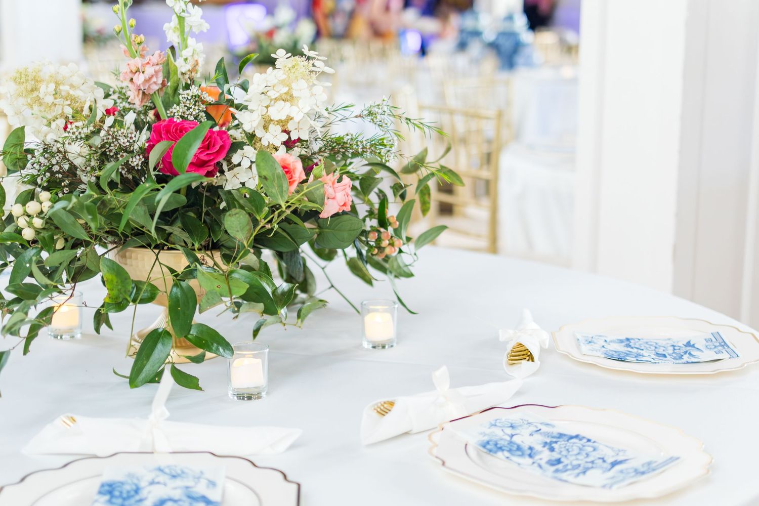 Elegant floral centerpiece with pink and white flowers on a white tablecloth with blue and white china place settings.