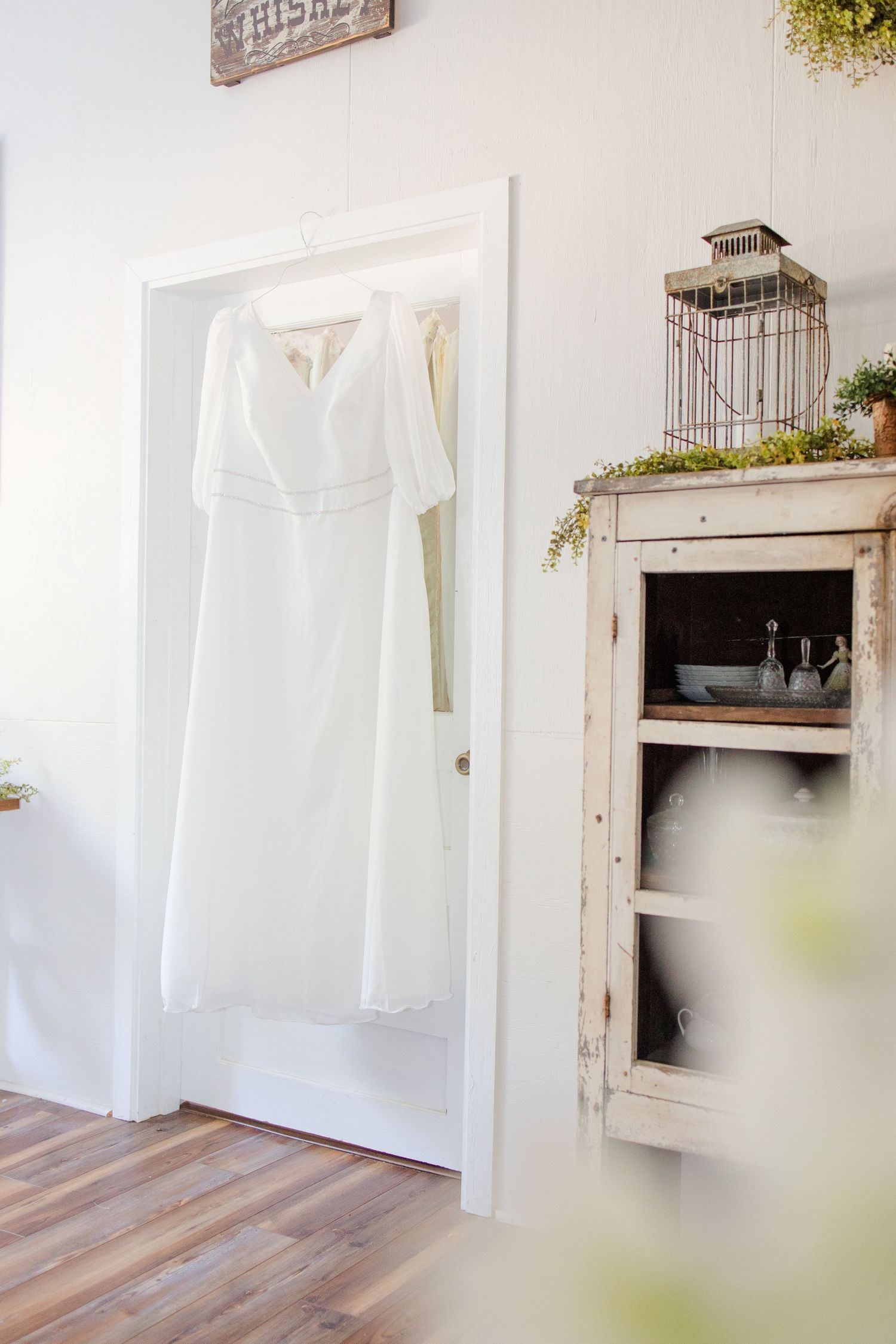 White wedding dress displayed on wall mount next to vintage cabinet with birdcage decor in rustic room setting.