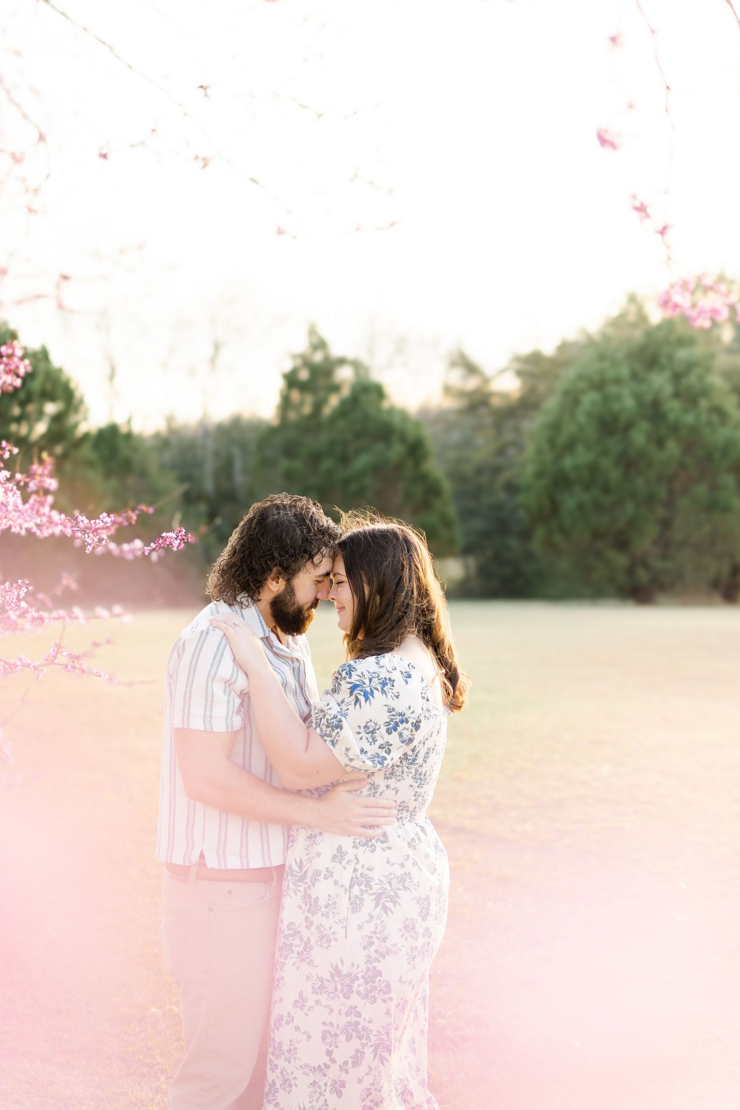 A romantic outdoor engagement photo session showing a couple embracing in a dreamy pink-tinted field at sunset.