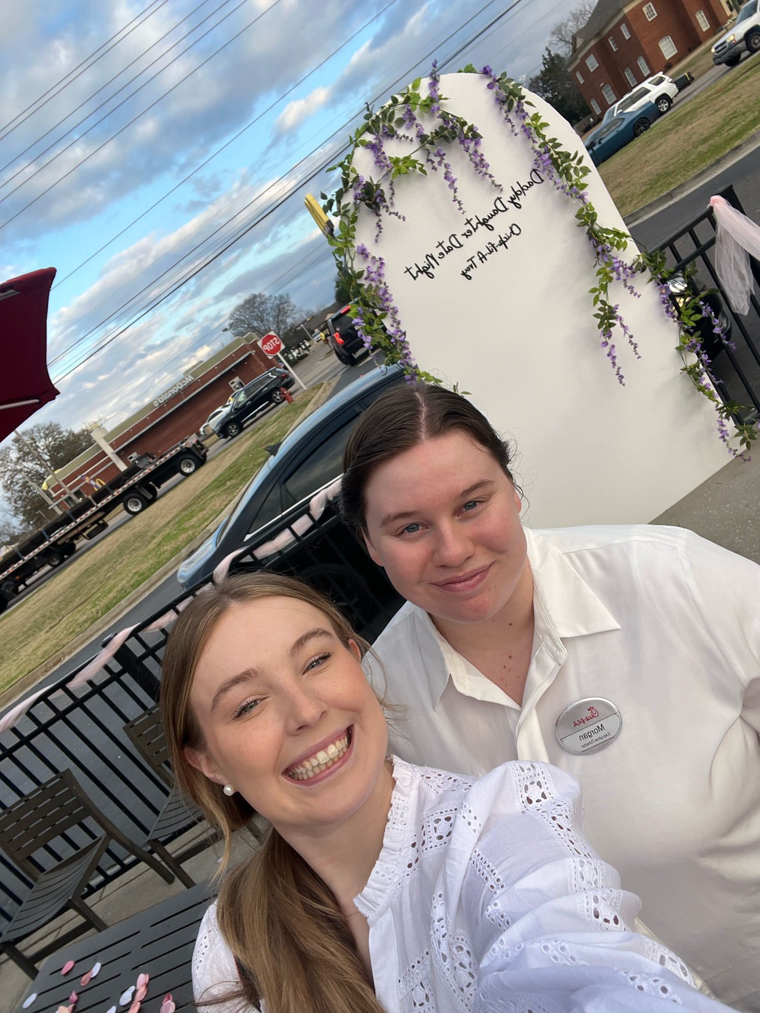 A couple takes a series of selfies outdoors on a sunny day with decorative floral arrangements in the background.