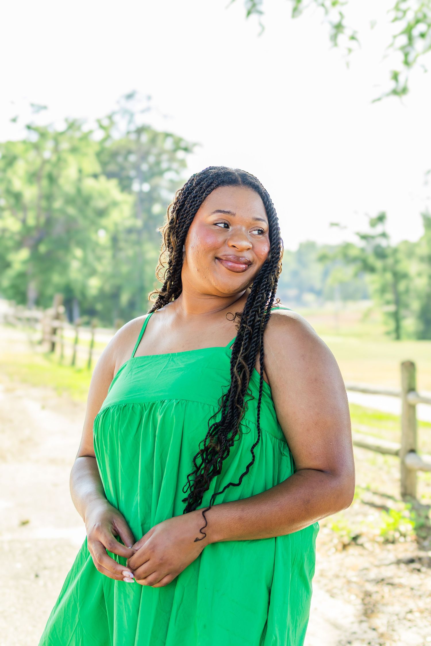 A woman in a flowing green dress poses outdoors in a sunny natural setting with a rustic wooden fence.