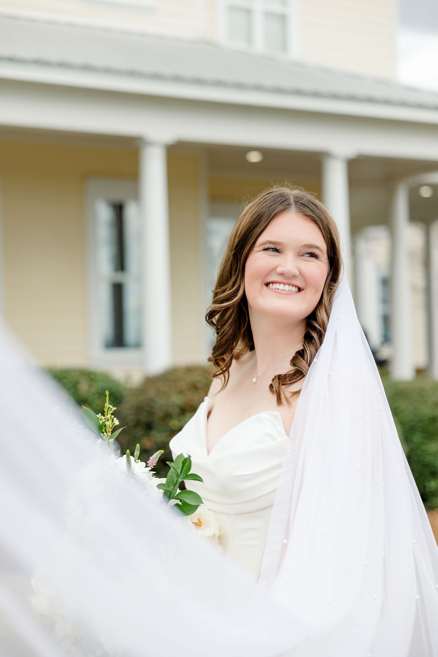 A bride in a white strapless wedding dress and flowing veil smiles brightly against a light colored building with columns.