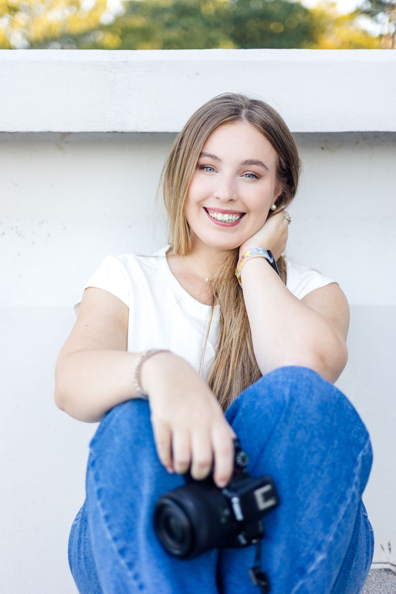 A person in blue jeans and white top sits casually while smiling at the camera.