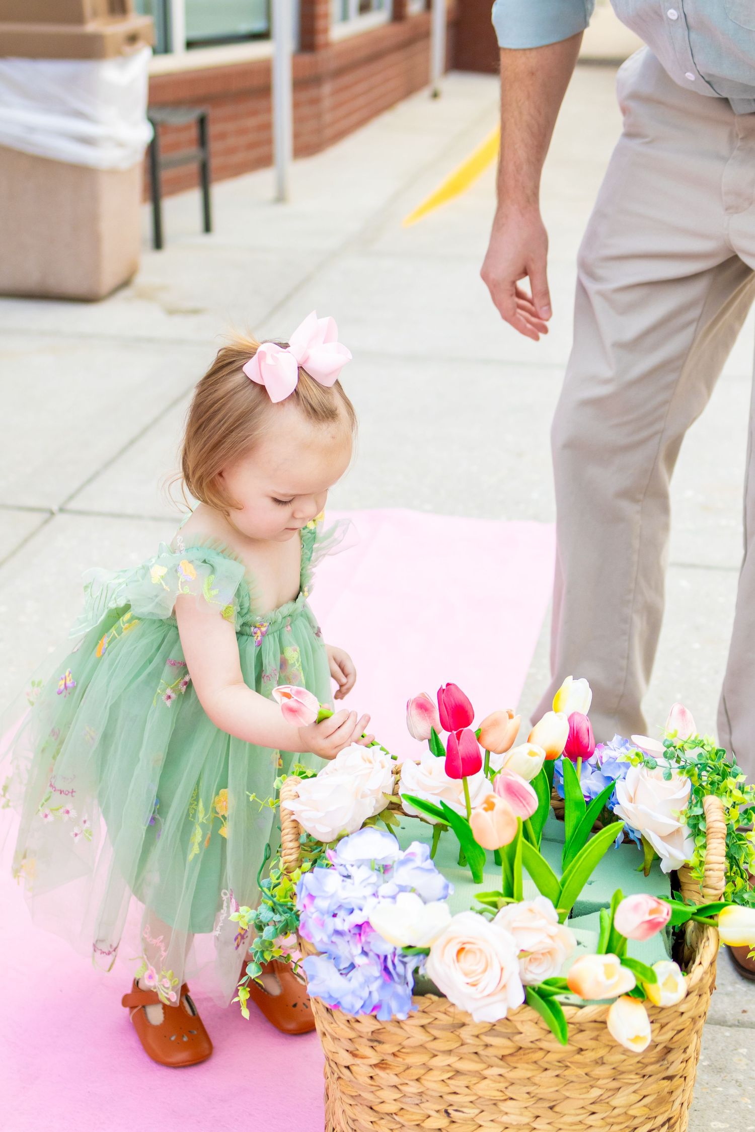 Small child in a mint green tulle dress reaching into a woven basket filled with colorful spring flowers.