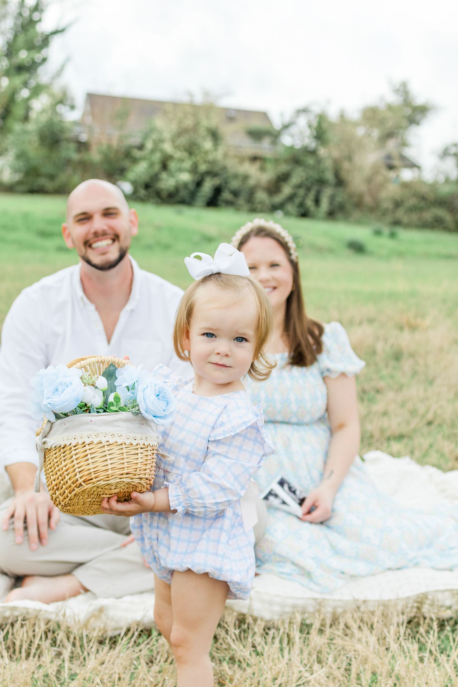 A family lounges together on a white blanket in a grassy meadow during a sunny summer day photoshoot.