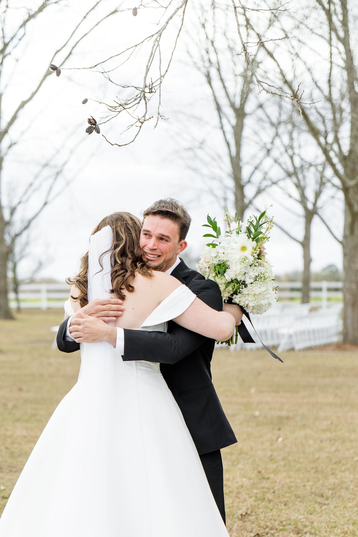 A couple shares a tender embrace during their wedding photoshoot in a winter field with bare trees in the background.