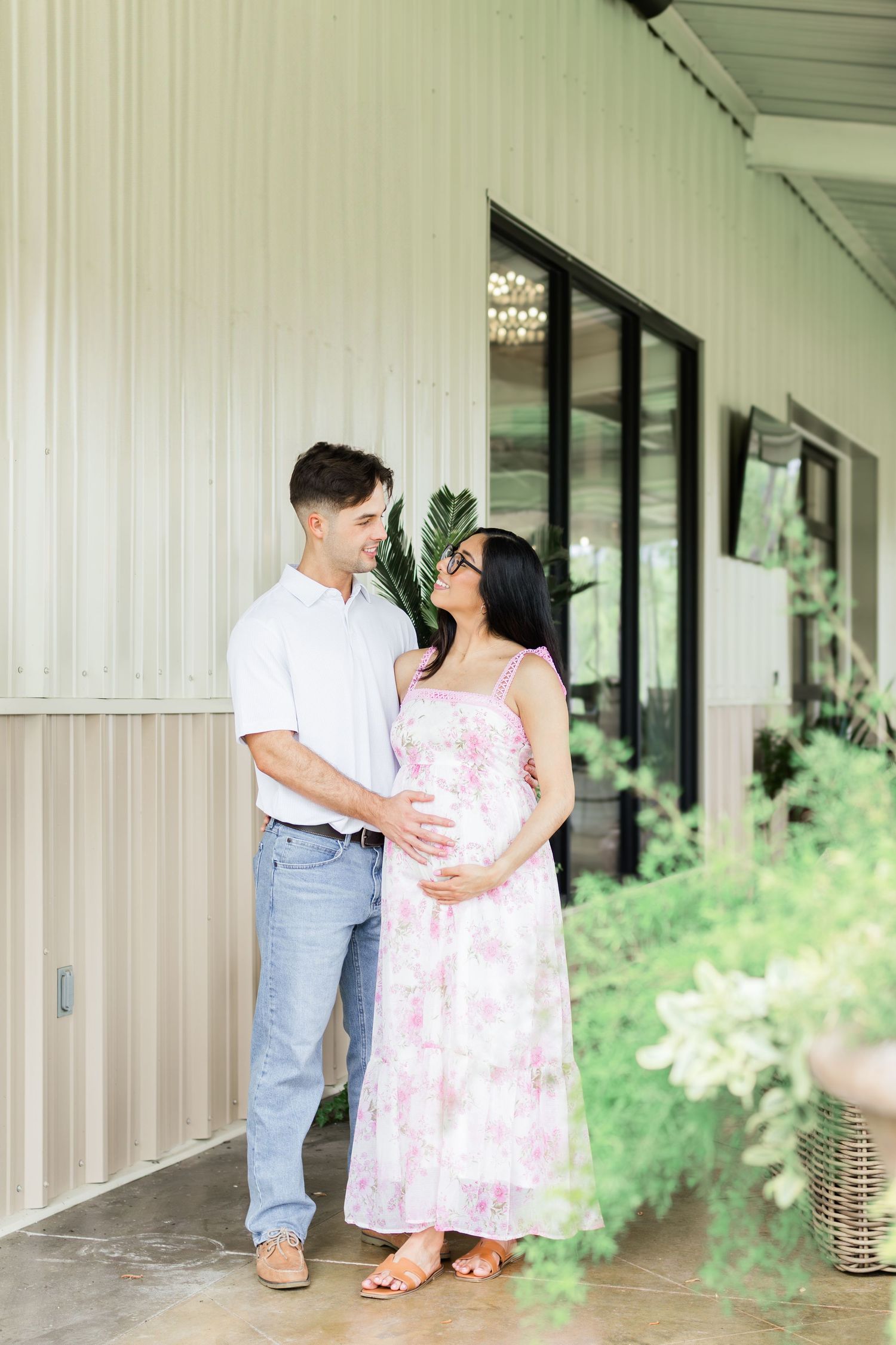 A couple stands together on a covered porch, with the expectant mother wearing a pink floral dress.