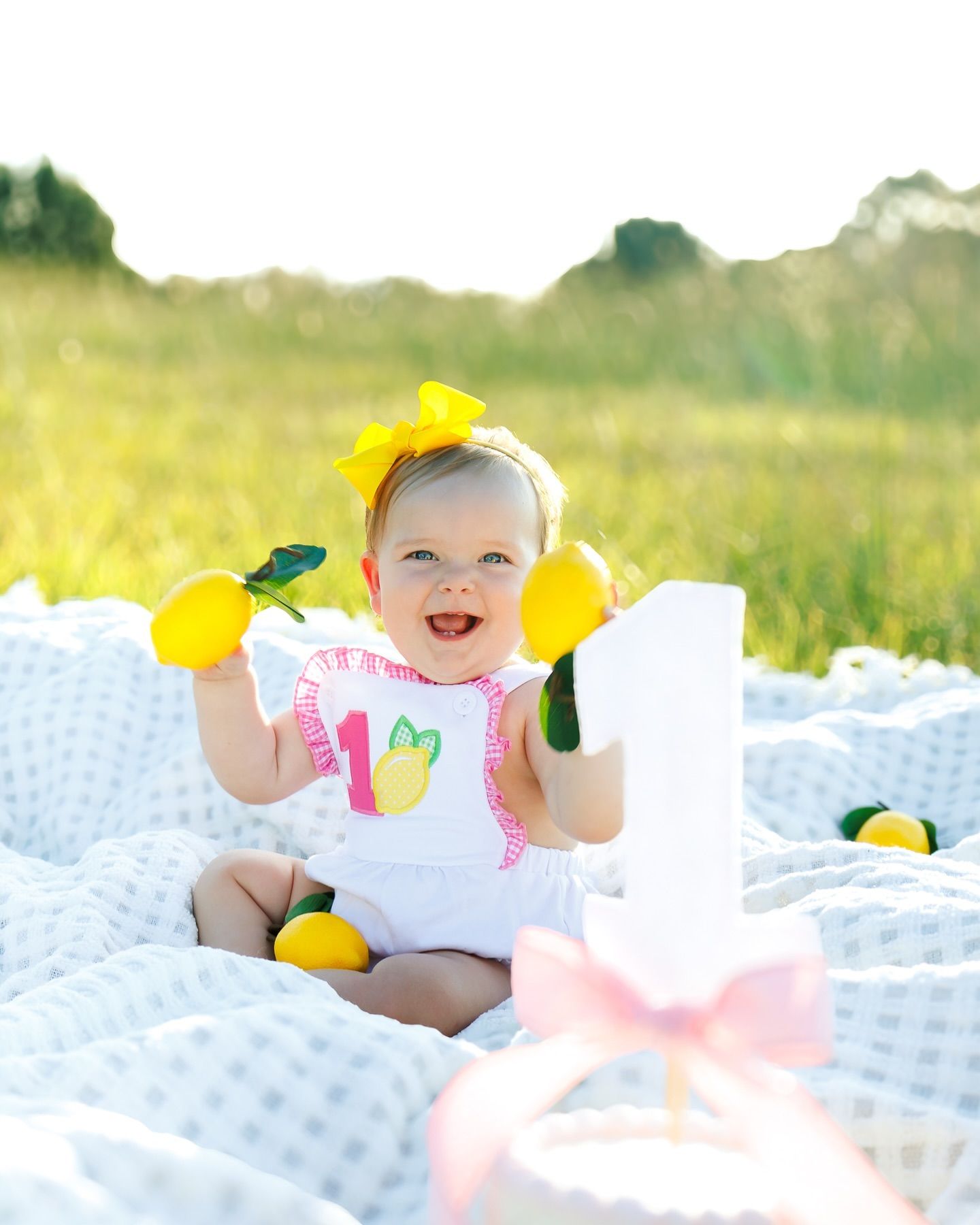 A smiling baby sits on a white blanket with yellow lemons and a matching yellow bow.