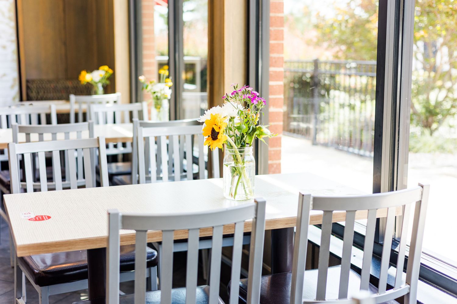 Modern restaurant interior with white chairs and tables arranged near large windows with yellow flower centerpieces.