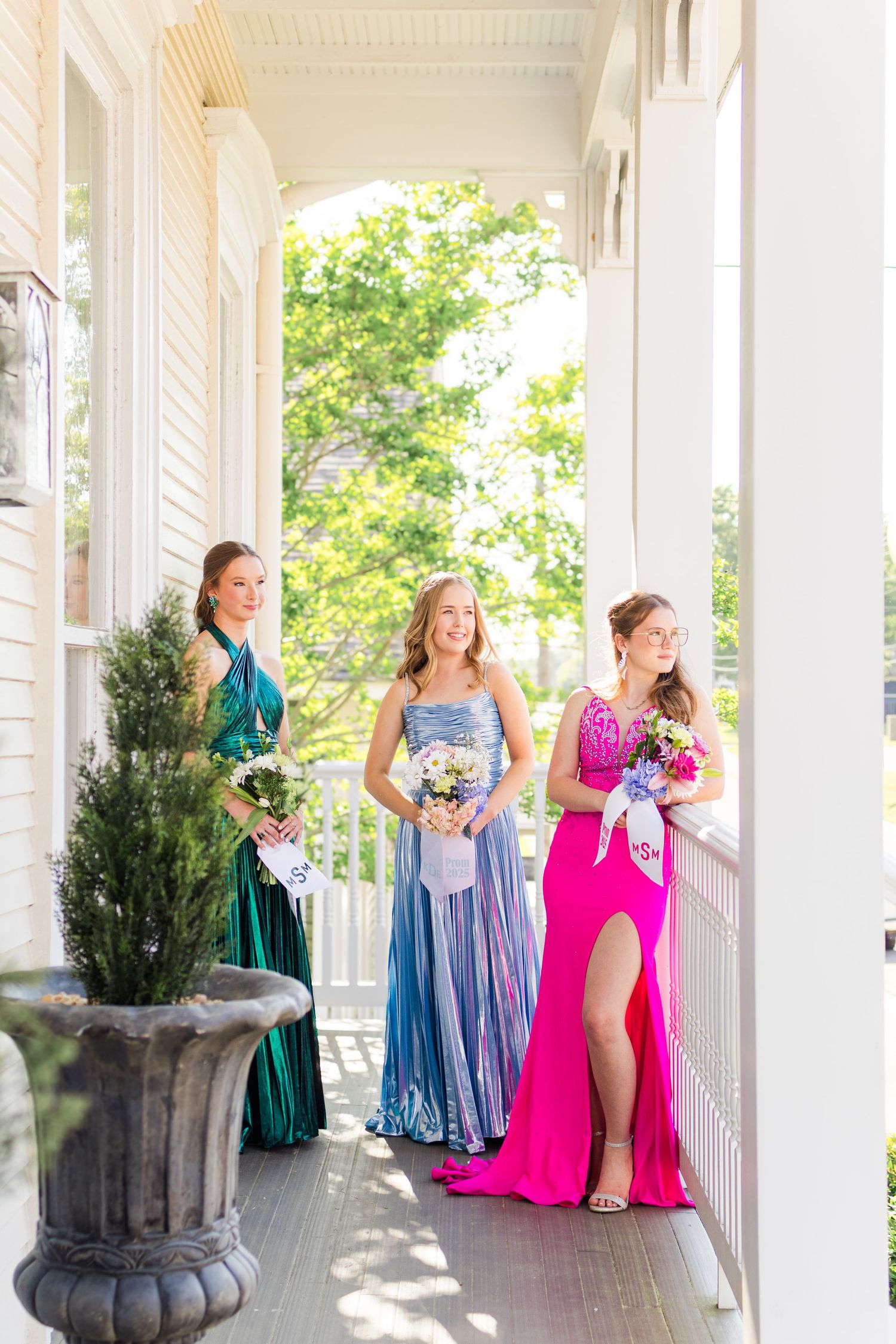 Bridesmaids in colorful formal dresses stand along a white wraparound porch adorned with tall planters.