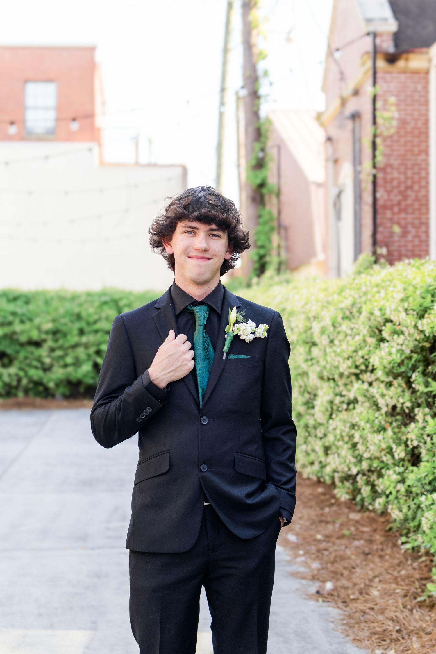 Young person in a black formal suit with boutonniere poses in front of brick building and landscaping.