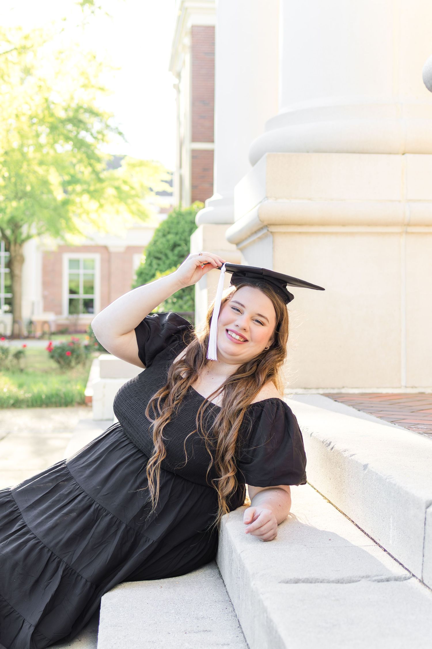 A graduate in a black dress and cap poses while sitting on a white stone ledge during a sunny outdoor photo session.
