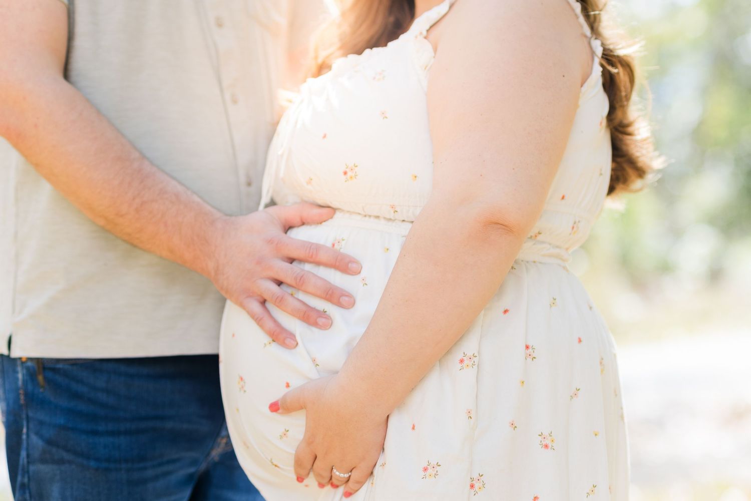Close up of expecting couple holding hands on pregnant belly in white dress and denim outdoors.