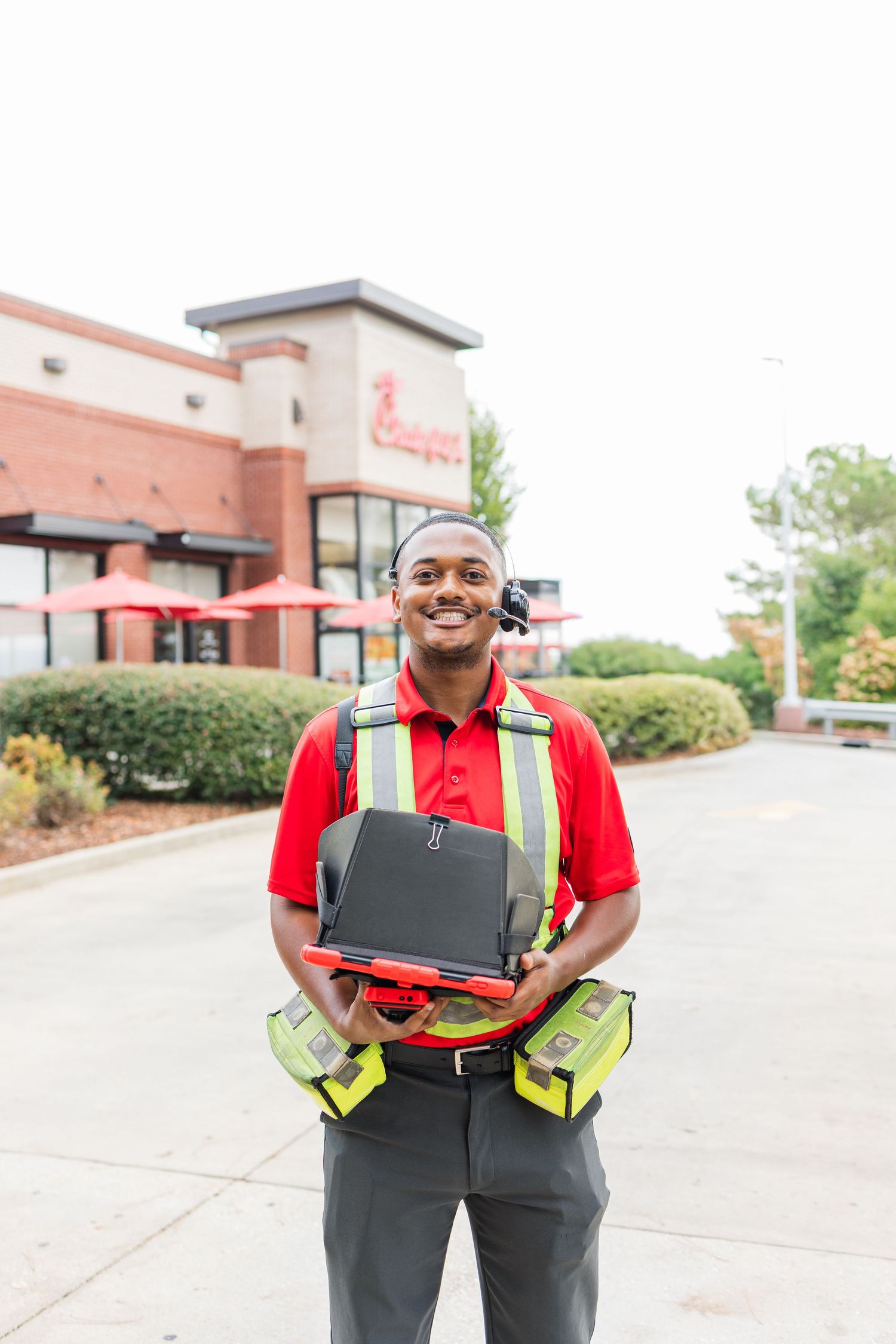 Worker in red polo shirt and safety vest holds laptop computer in parking lot outside commercial building.