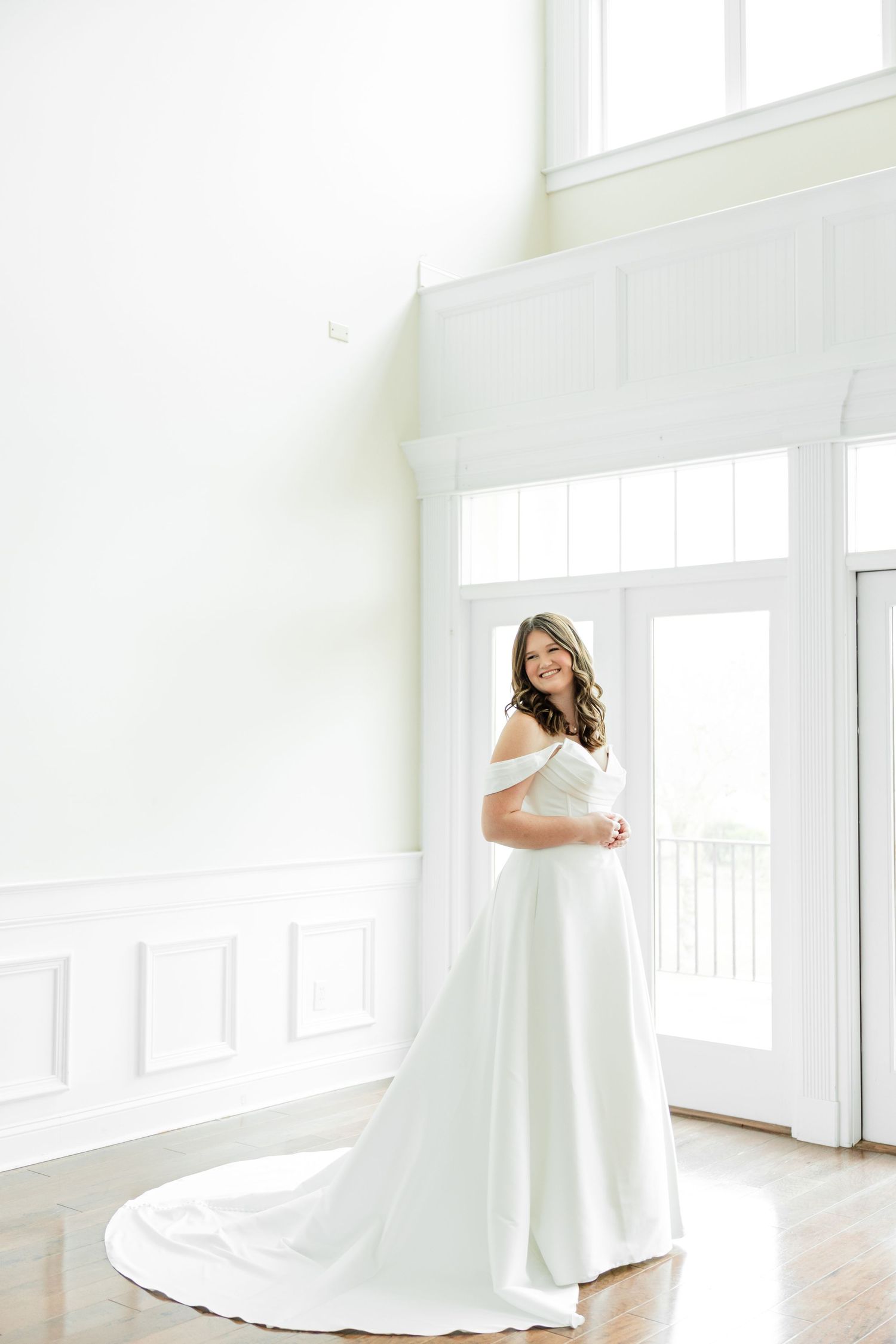 A person in an elegant off-shoulder white wedding gown stands in a bright, minimalist room with white walls and wood floors.