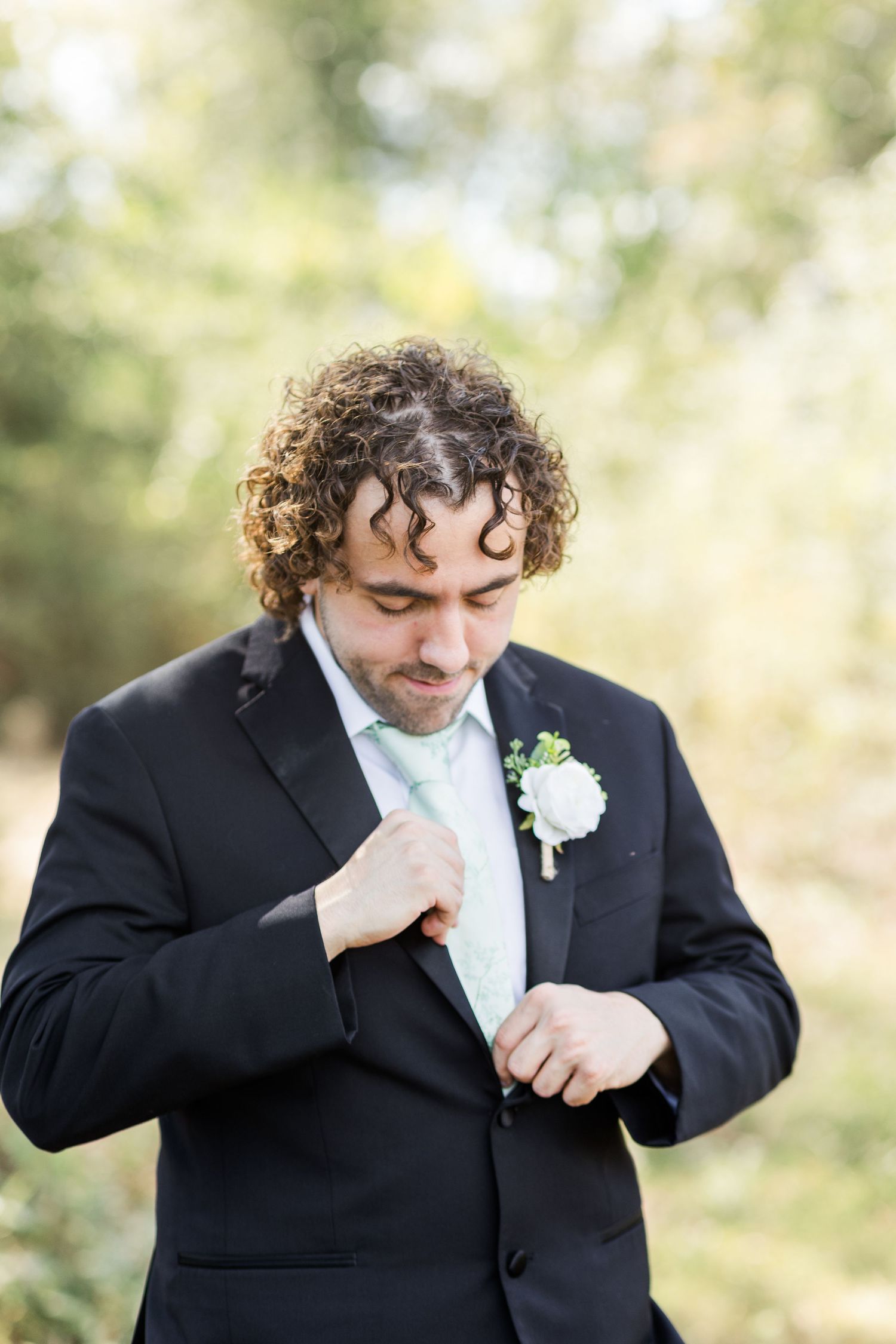 A person in a black tuxedo and white bowtie adjusts their lapel while wearing a white boutonniere.