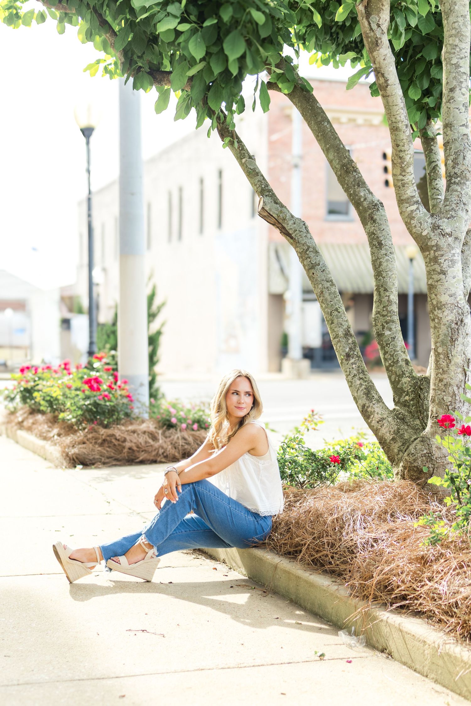 Person in casual wear leans against textured brick wall next to flowering plants and trees.