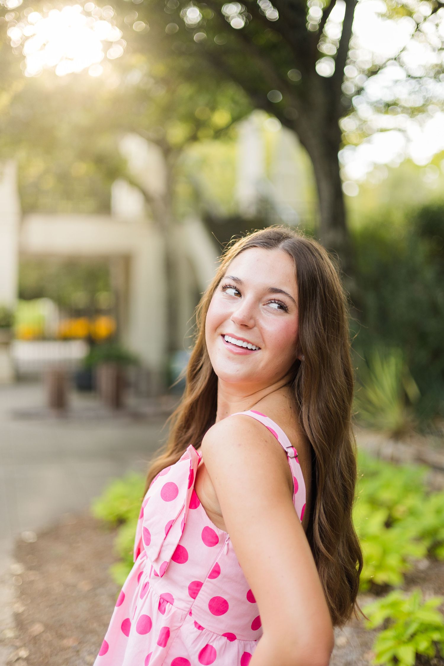 A person in a pink polka dot dress smiles while posing outdoors in a garden setting with blurred trees in the background.