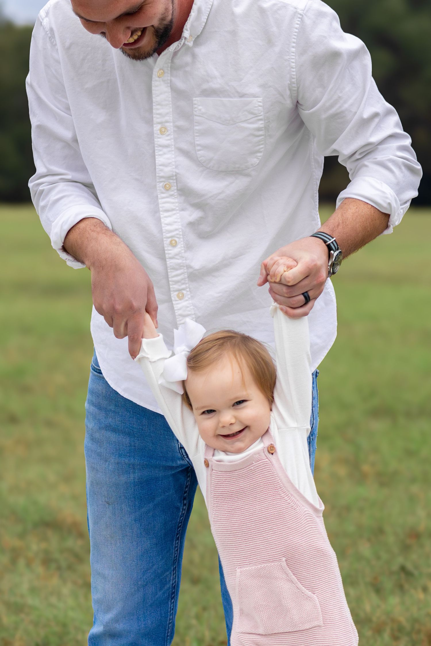 An adult in a white button-down shirt and jeans plays with a toddler wearing a pink dress in a grassy field.