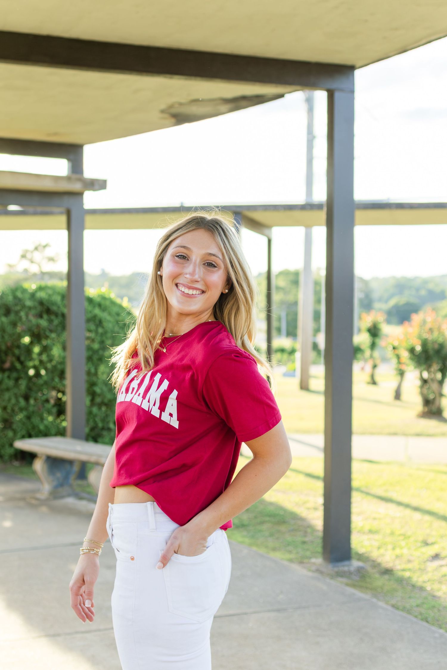 Someone wearing a red Alabama t-shirt and white pants poses outdoors under a covered patio area on a sunny day.