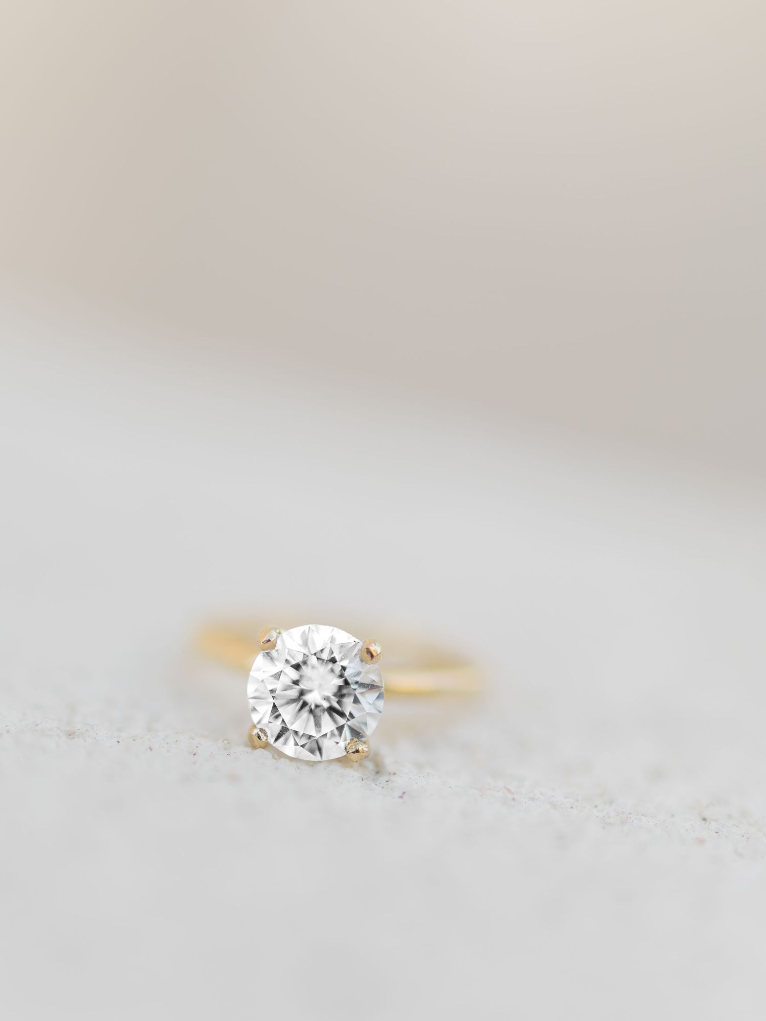 Gold solitaire engagement ring with round brilliant diamond displayed on a textured light gray surface.