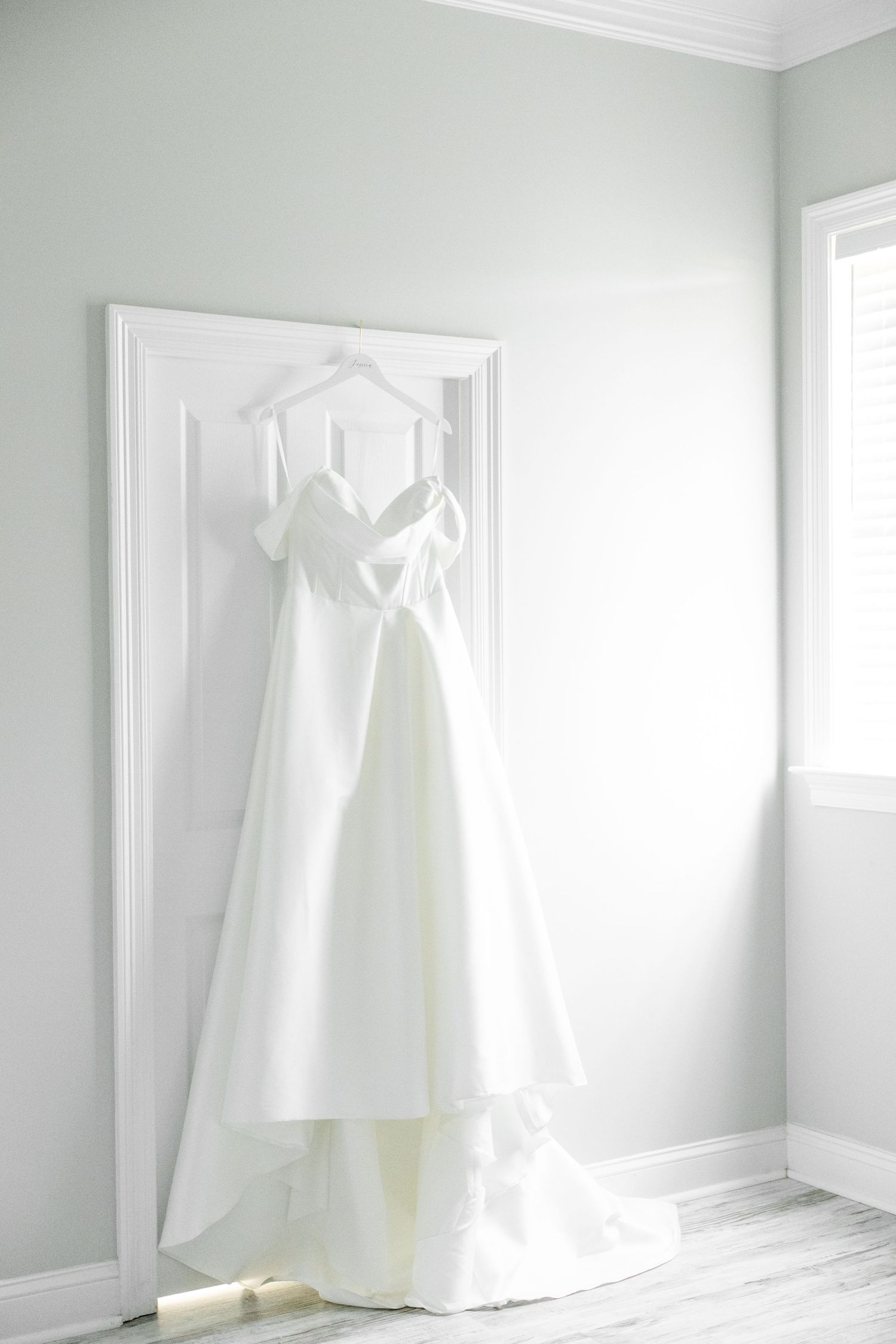 Elegant white wedding dress with bow detail hanging on a door in a soft, light-filled bridal suite.