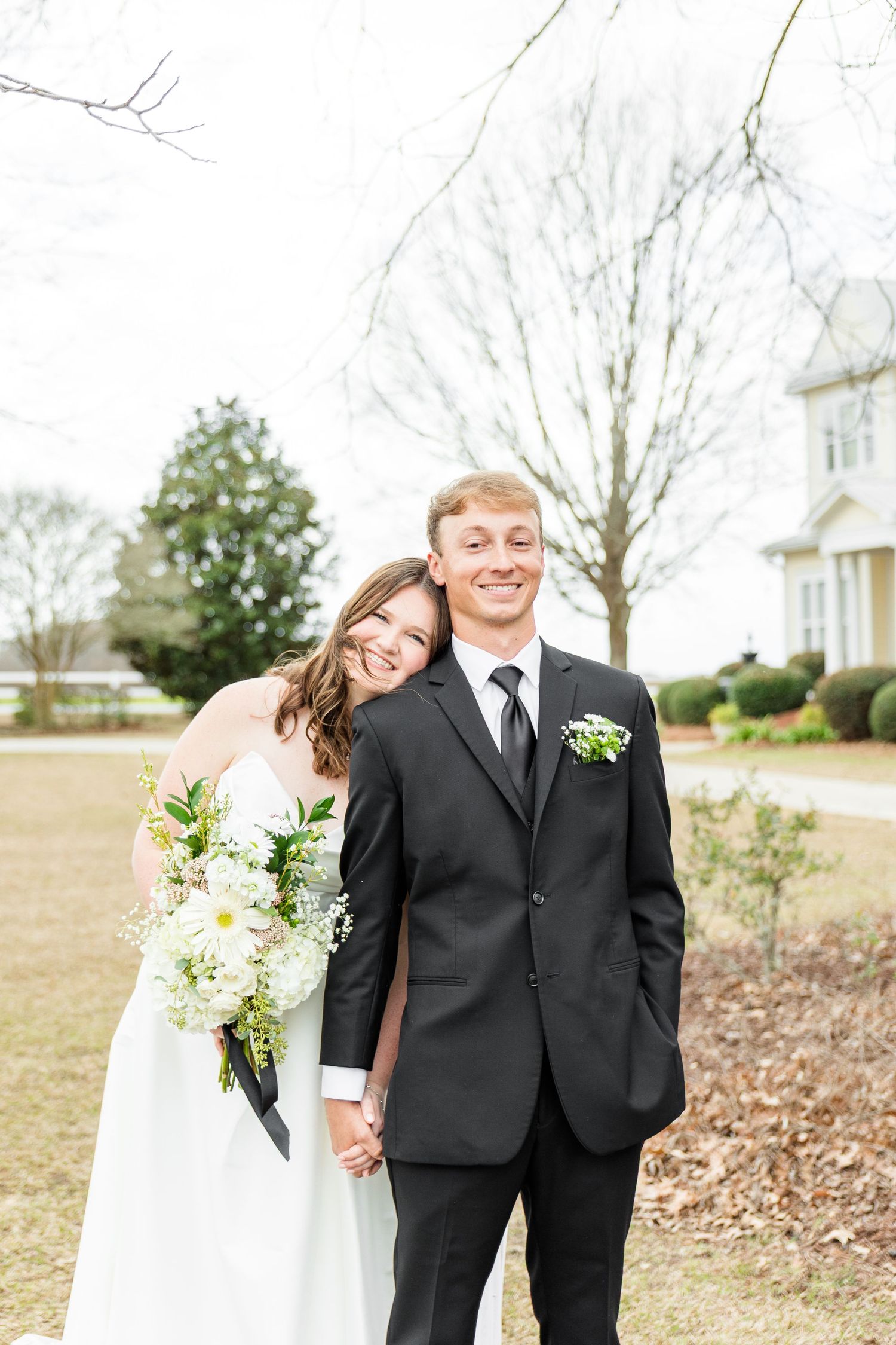 A joyful wedding couple shares a candid moment outdoors with white flowers and greenery in the background.